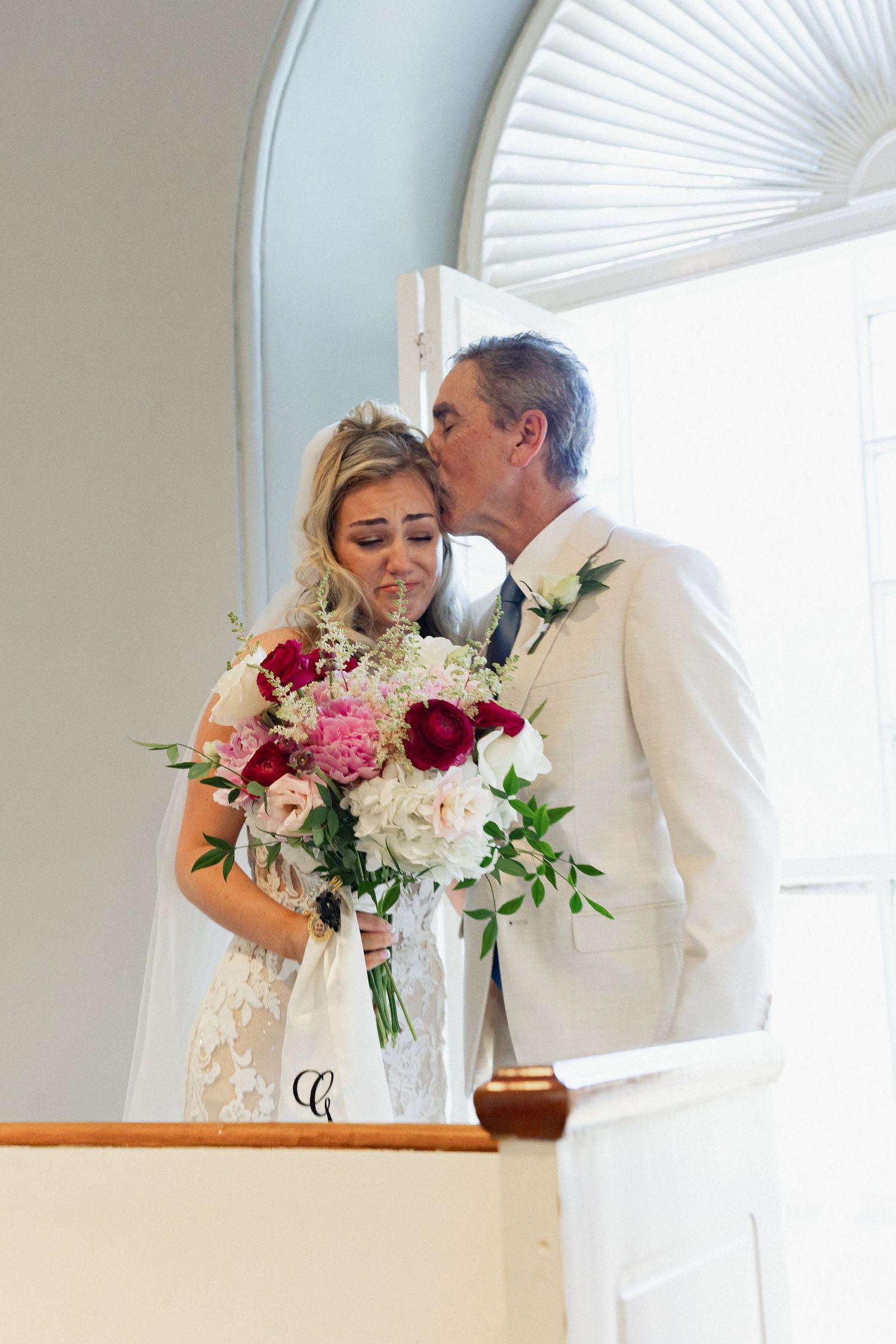 Father of the bride kisses his emotional daughter holding a colorful bouquet of red and pink roses on her wedding day.