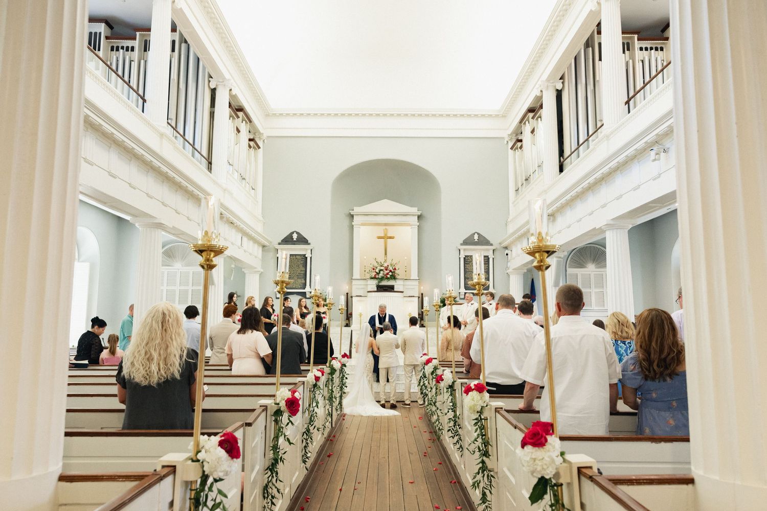 Wide angle view of elegant church wedding ceremony with floral-decorated pews and guests seated.
