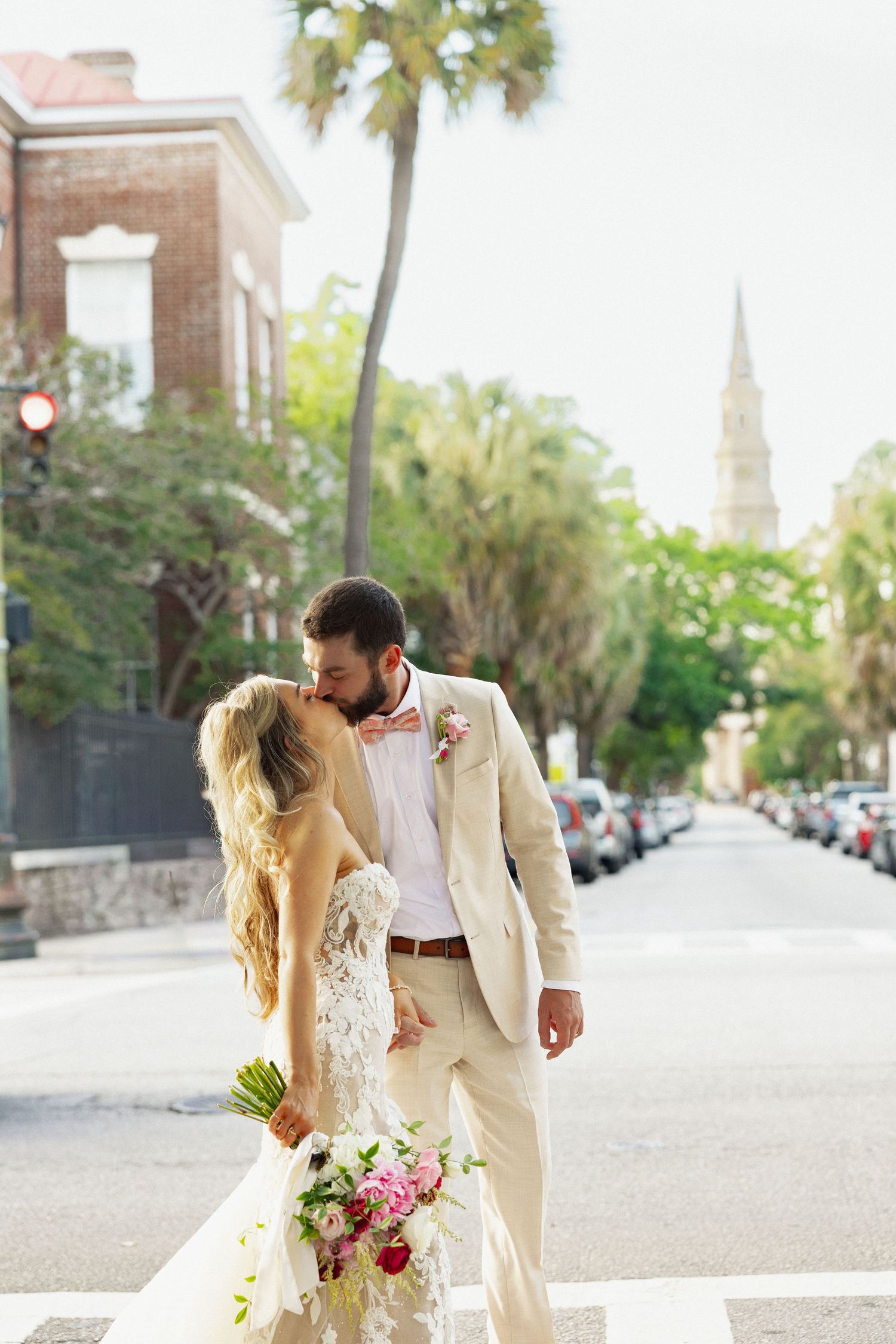 Bride and groom share romantic kiss on Charleston street crossing with church steeple and palm trees behind them.