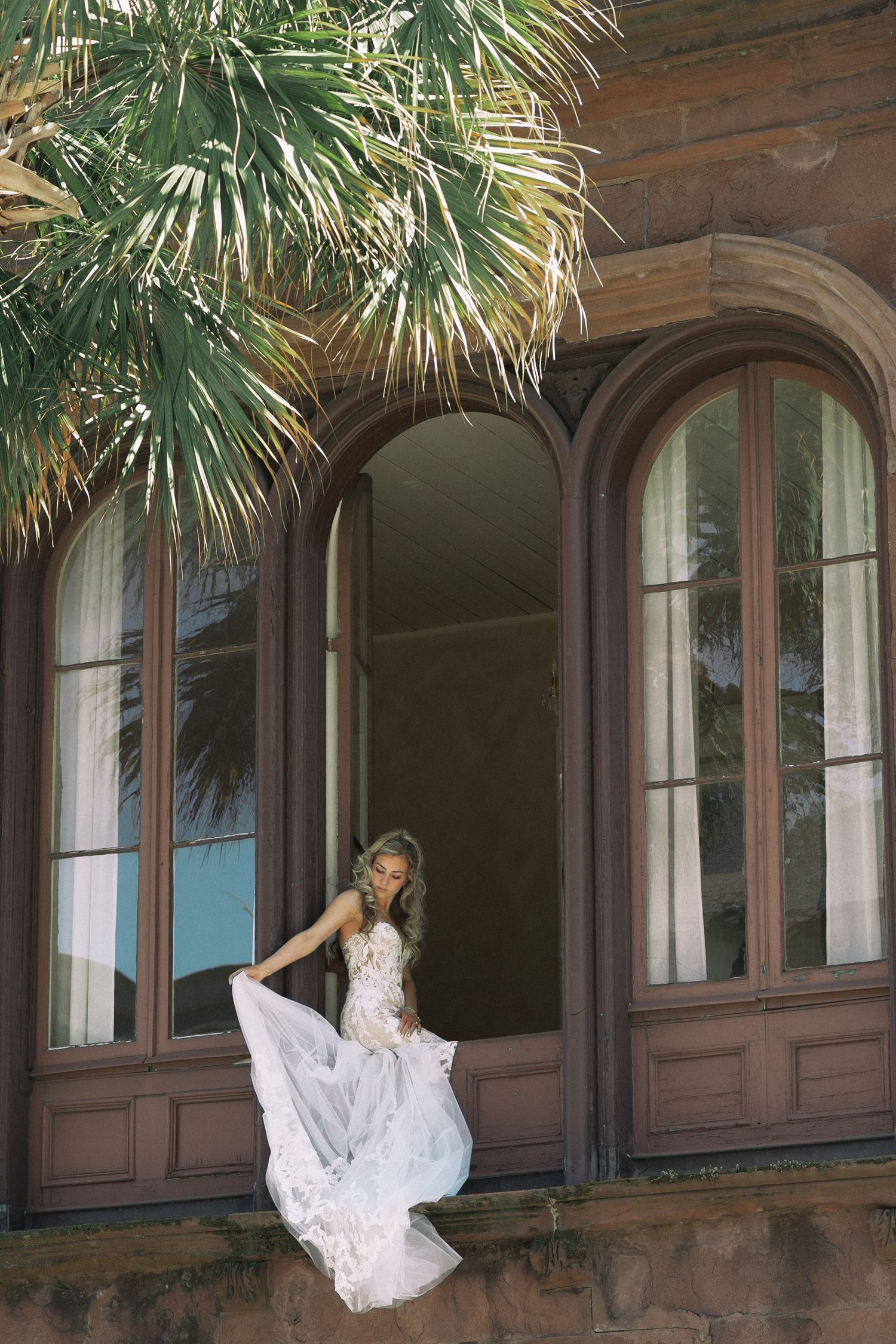 Bride in lace gown poses on historic building window ledge with flowing skirt and palm tree backdrop.