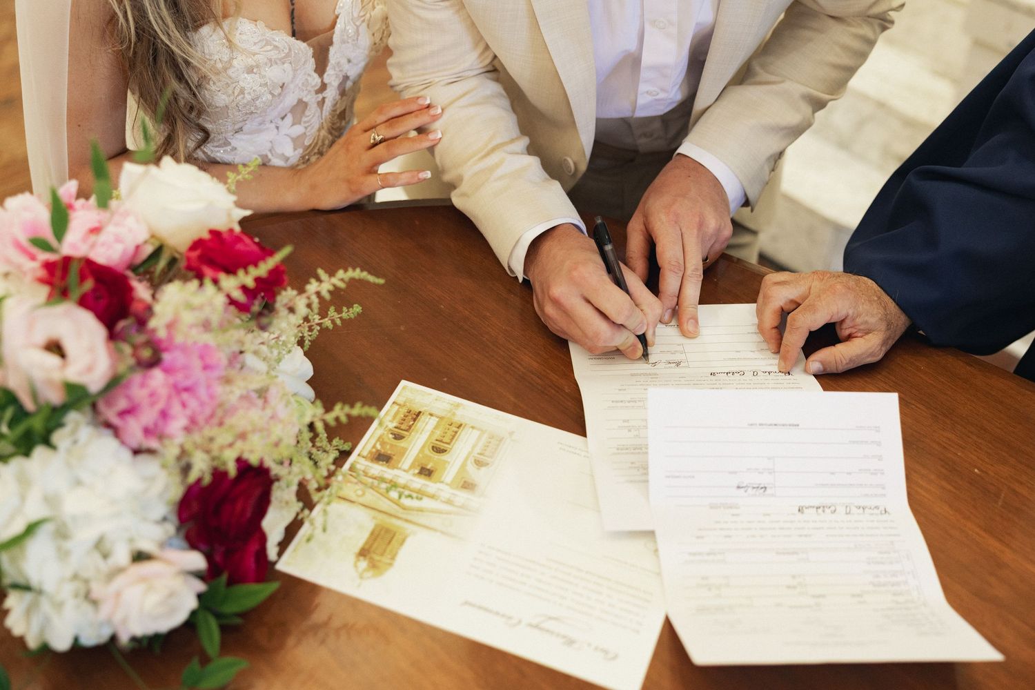 Bride and groom signing wedding documents at a wooden table with a bouquet of pink and red flowers nearby.