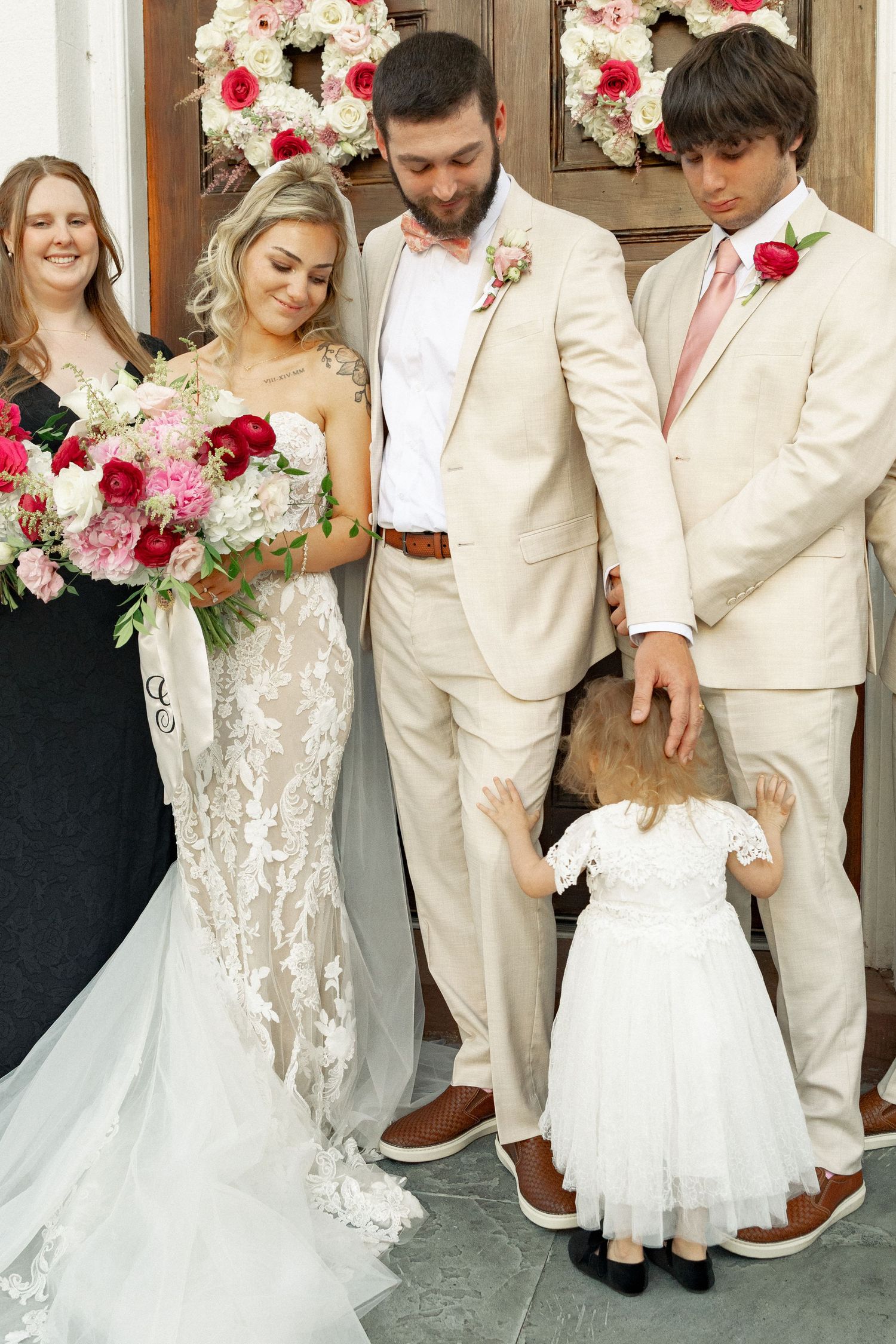 Bride in lace gown holds pink bouquet with groom in tan suit, groomsman, flower girl, and bridesmaid at wedding.