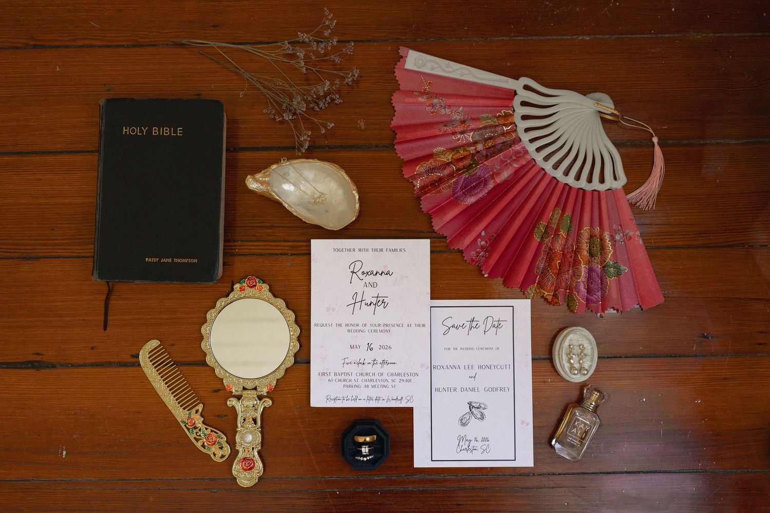 Wedding flat lay with Holy Bible, pink fan, invitation suite, vintage mirror, comb, and perfume on wood table.
