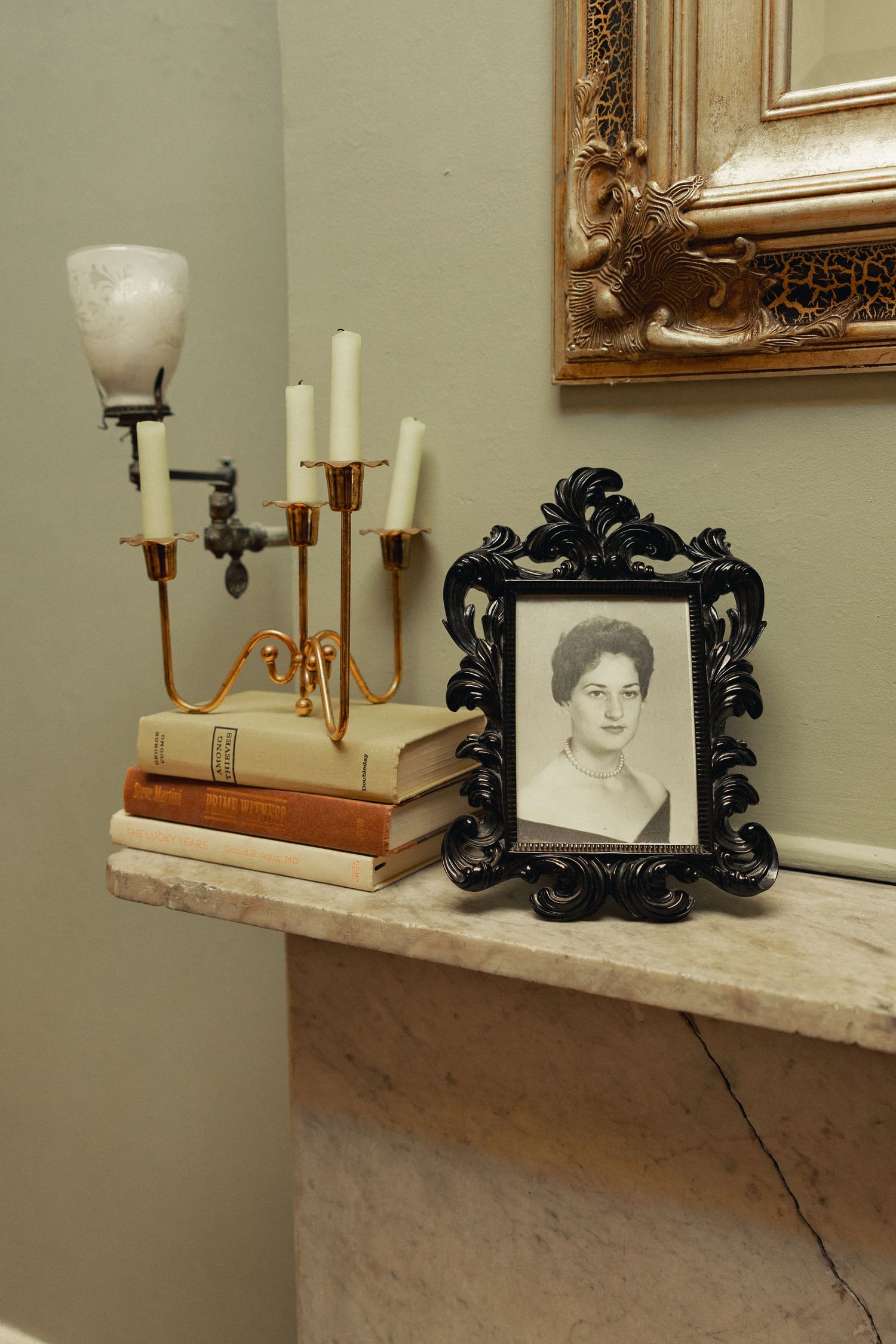 Vintage mantel display with black ornate framed portrait, gold candelabra, and stacked antique books on marble shelf.