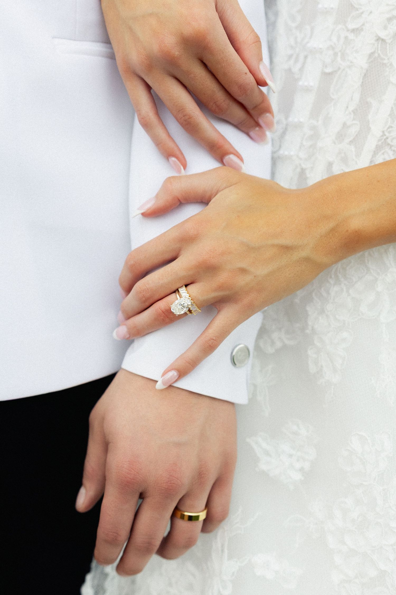 Bride and groom showing wedding rings, gold band and diamond engagement ring, hands resting on white suit jacket.