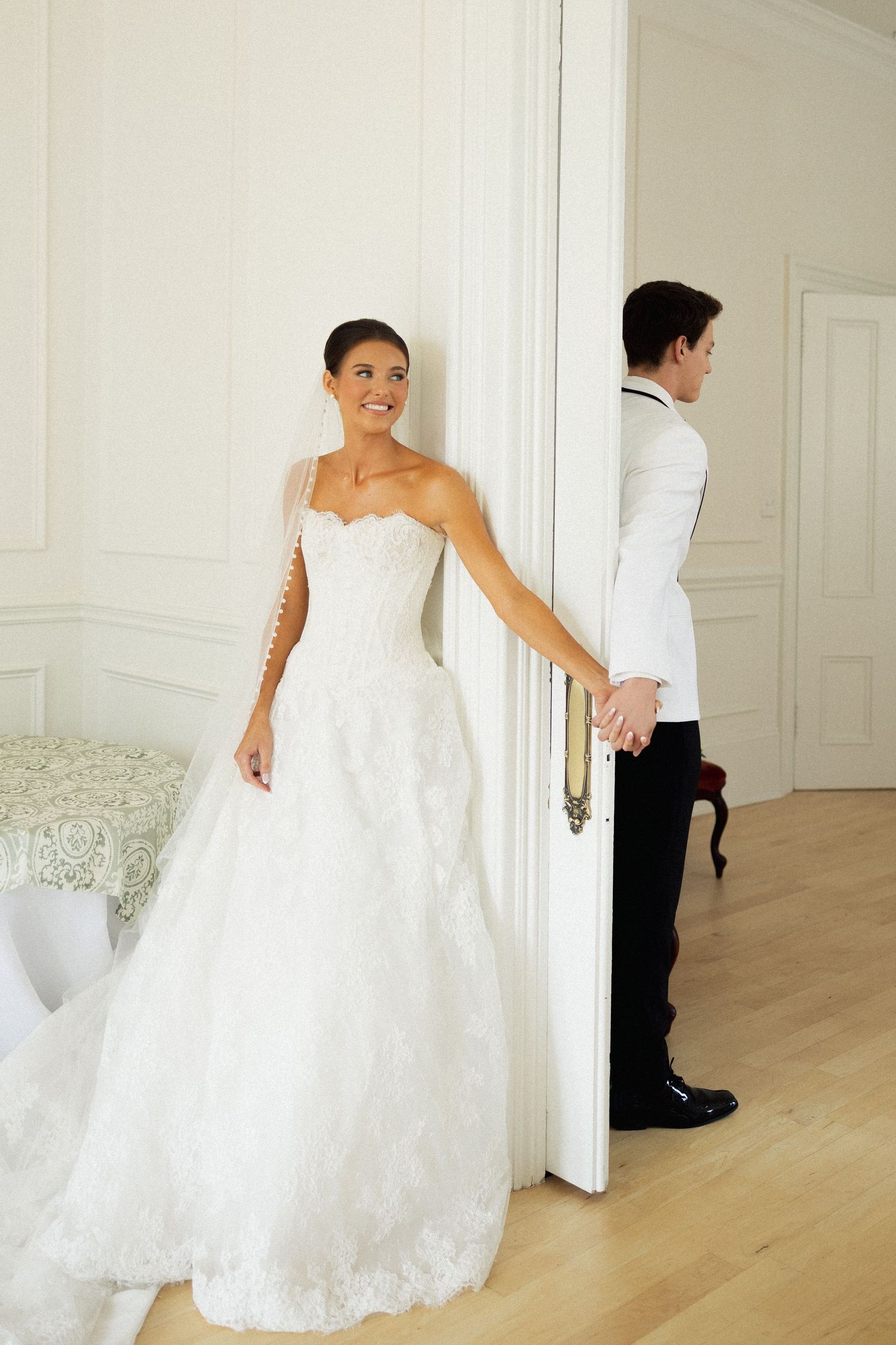 Bride in lace ball gown holds groom's hand around a door in a first look wedding moment.