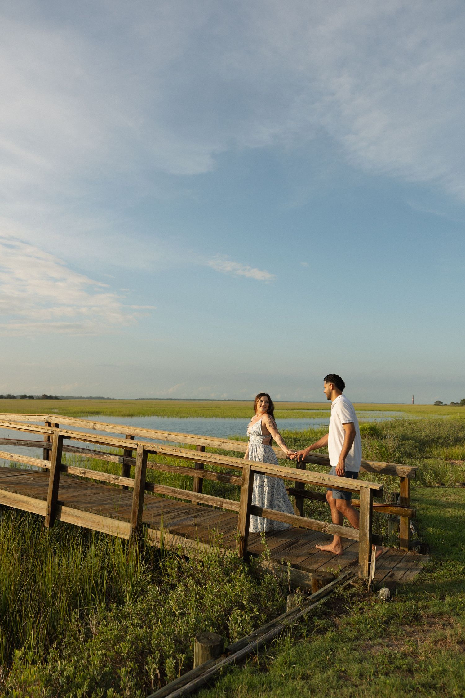 Couple holding hands on a wooden bridge overlooking a serene marsh and lake under a blue sky.