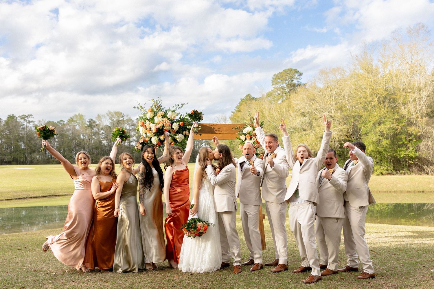 Joyful wedding party celebrating outdoors, bridesmaids in gold and rust gowns, groomsmen in tan suits, bride and groom center.