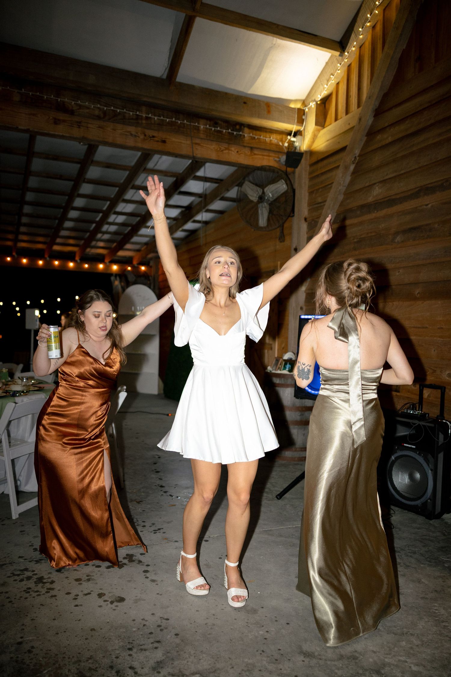 Bride in white mini dress dances joyfully with arms raised alongside bridesmaids at barn reception.