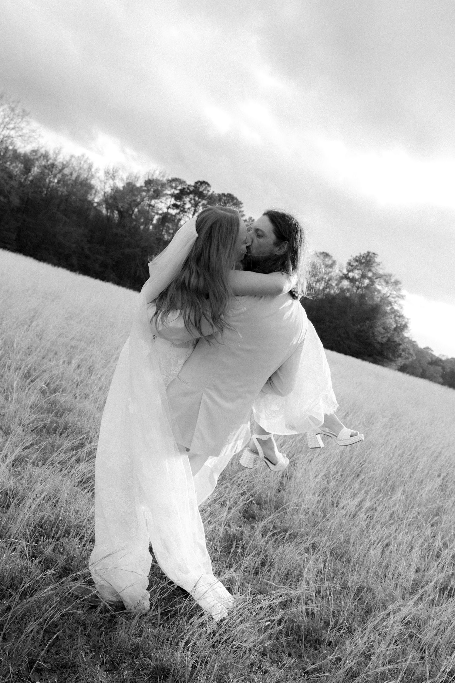Black and white photo of groom lifting and kissing bride in field under cloudy sky.