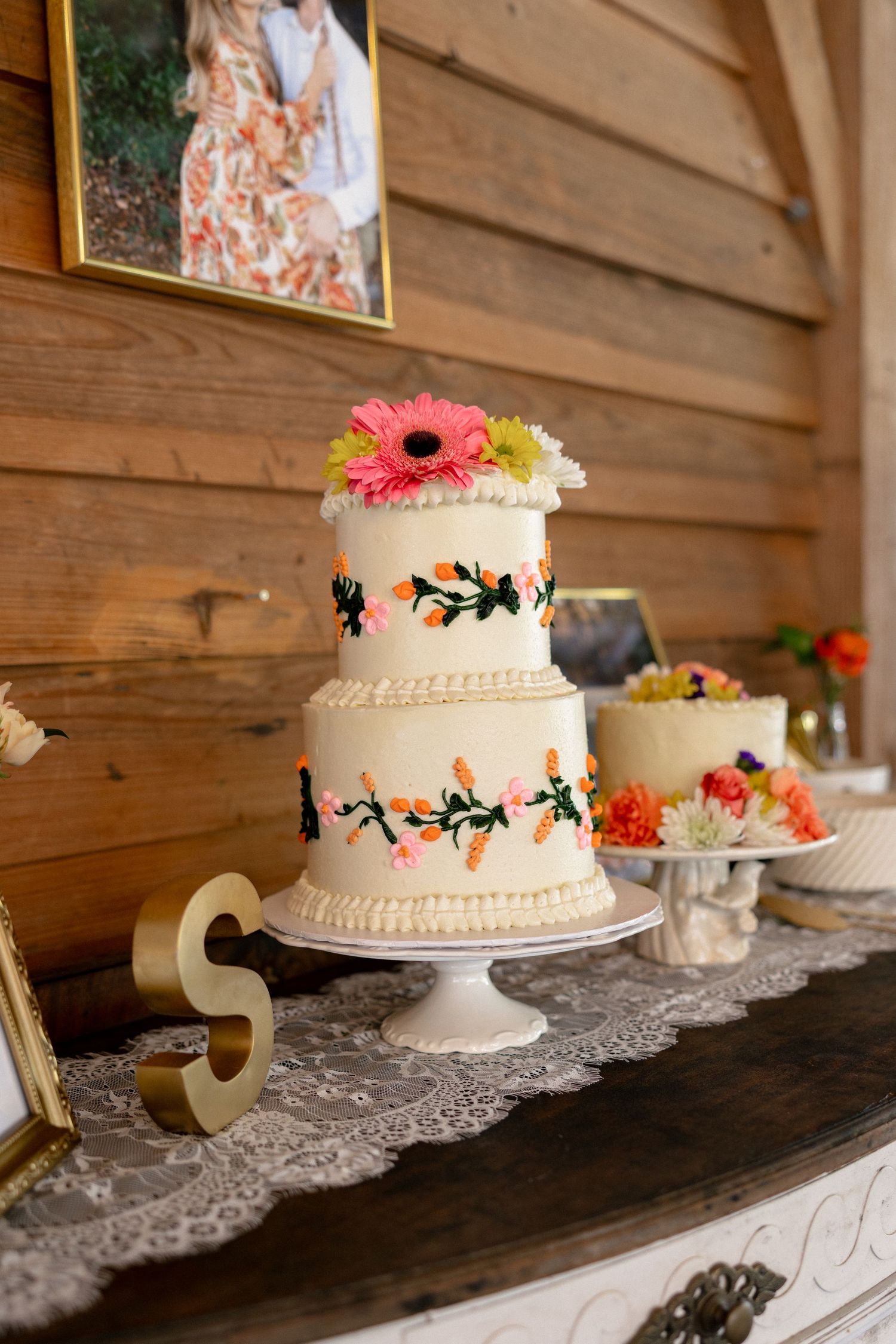 Two-tier white wedding cake decorated with colorful floral designs and fresh gerbera daisies on rustic wood backdrop.