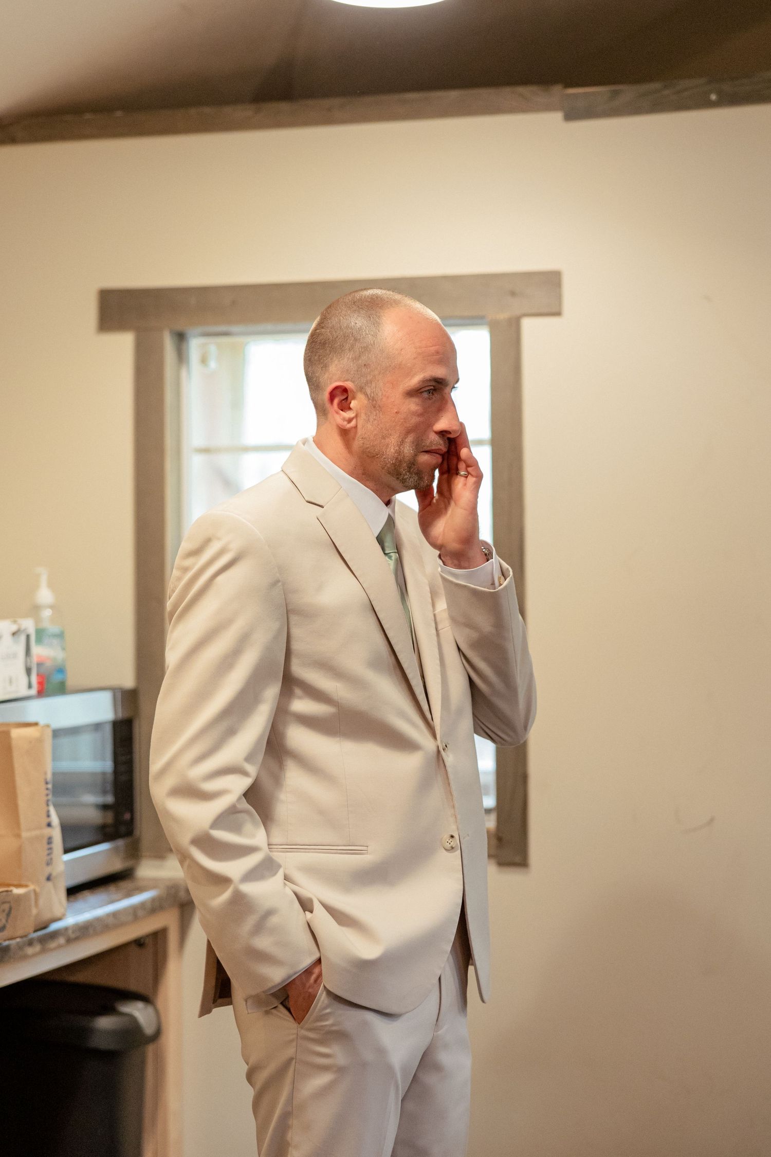 Bald groom in beige suit talks on phone while standing in a rustic wedding preparation room.