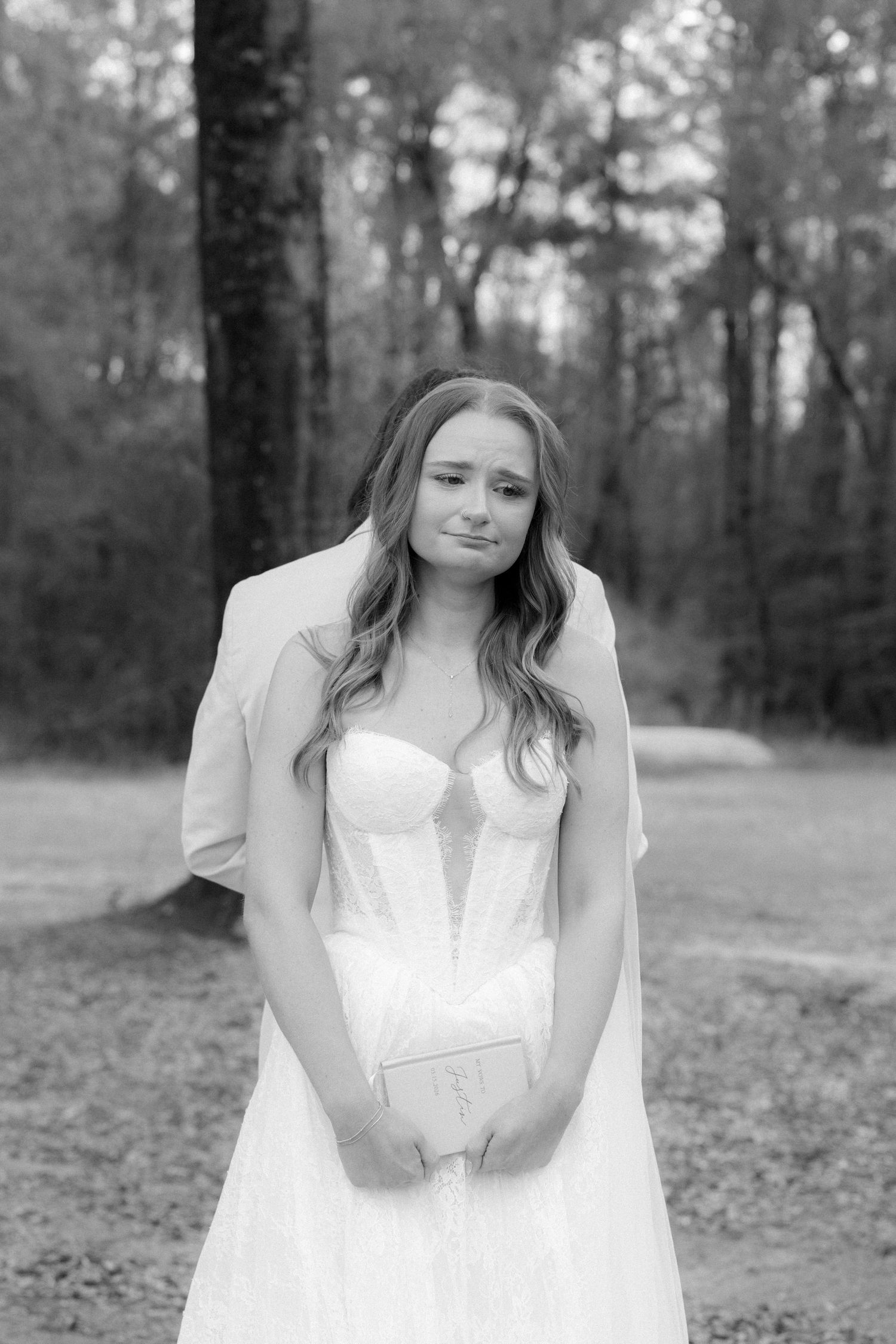 Black and white portrait of emotional bride in strapless lace gown holding vow book outdoors.