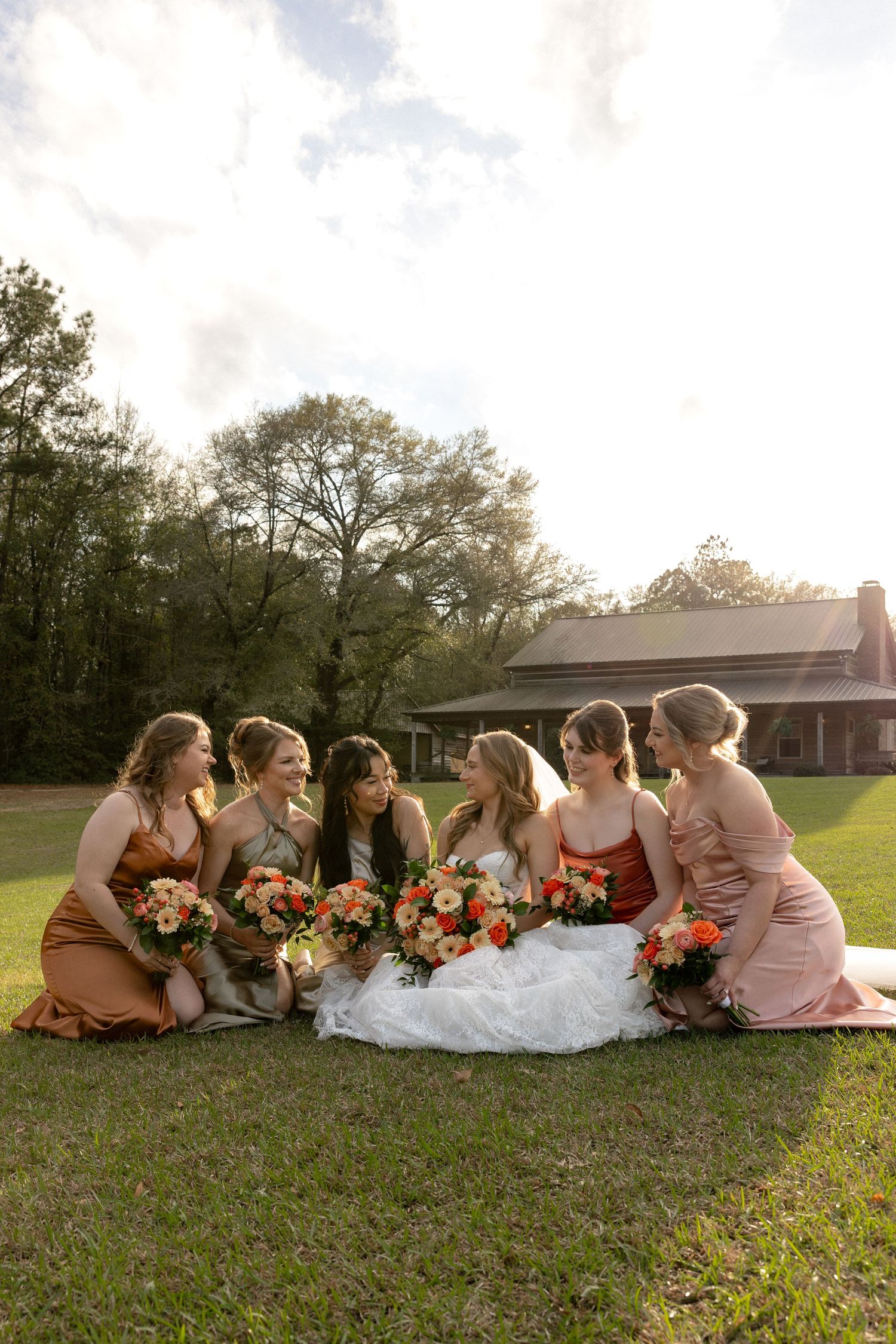 Bride and five bridesmaids sitting on grass laughing together in earth-tone satin dresses at outdoor venue.
