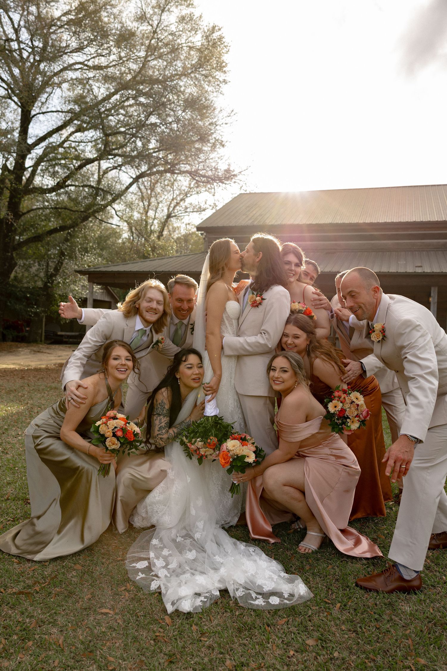 Joyful wedding party poses with kissing newlyweds in warm sunlight, holding colorful orange and pink bouquets.