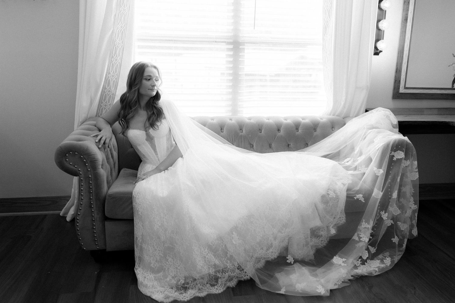 Black and white portrait of bride in lace gown and veil resting on elegant sofa.