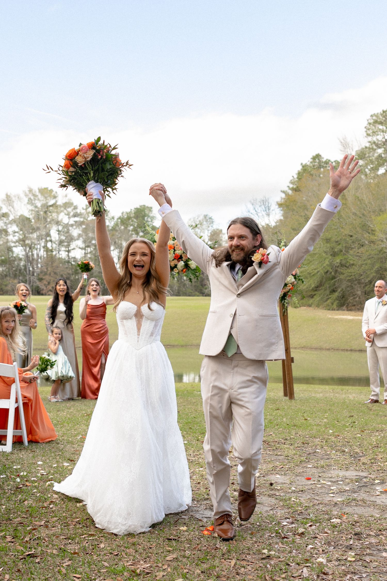 Joyful newlyweds raise hands walking down outdoor wedding aisle, bride in white gown holding orange bouquet.