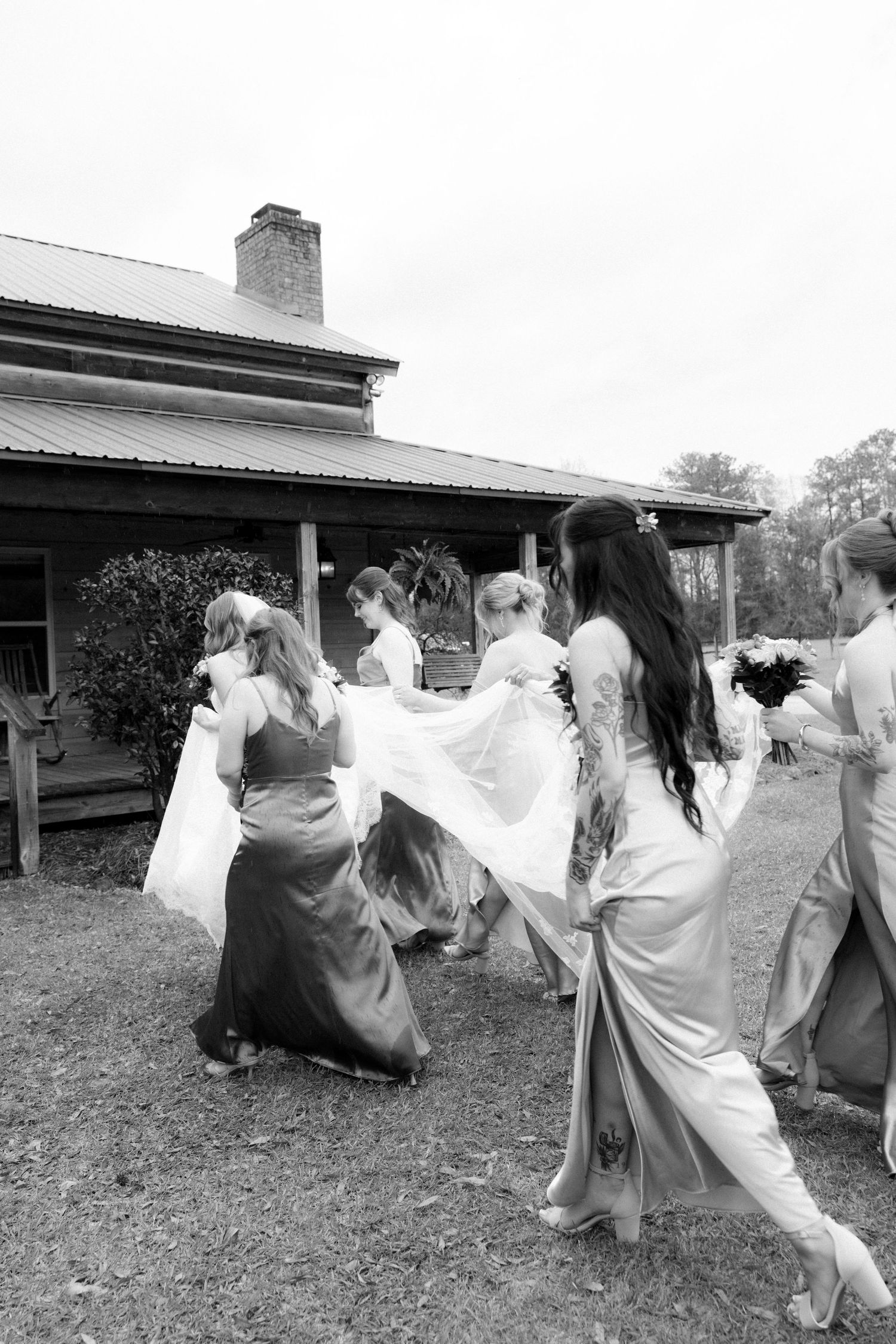 Wedding party walks toward rustic farmhouse as bridesmaids carry bride's flowing white dress train in black and white.