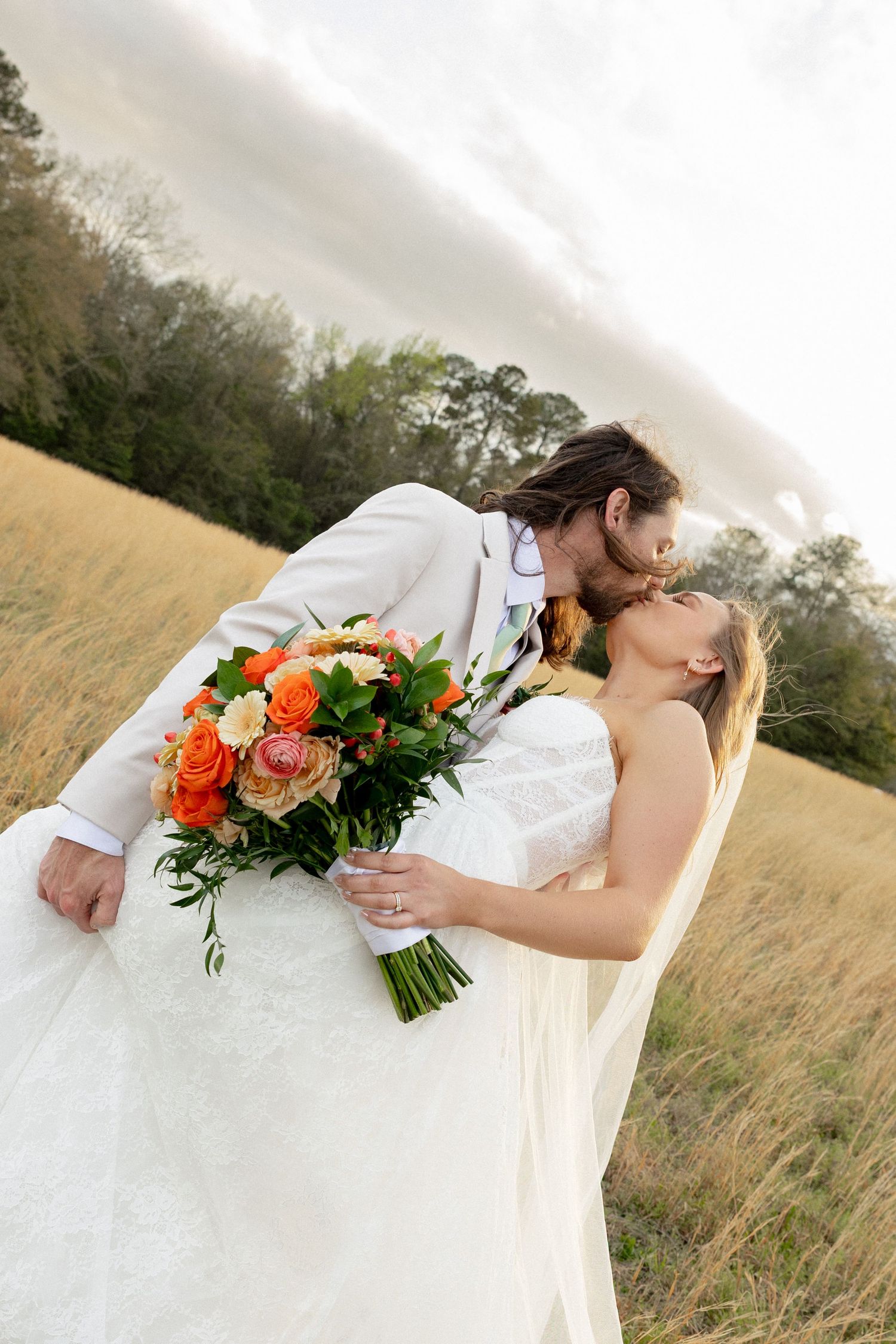 Bride and groom kissing in a golden meadow, bride holding vibrant orange and coral bouquet, groom in ivory suit.