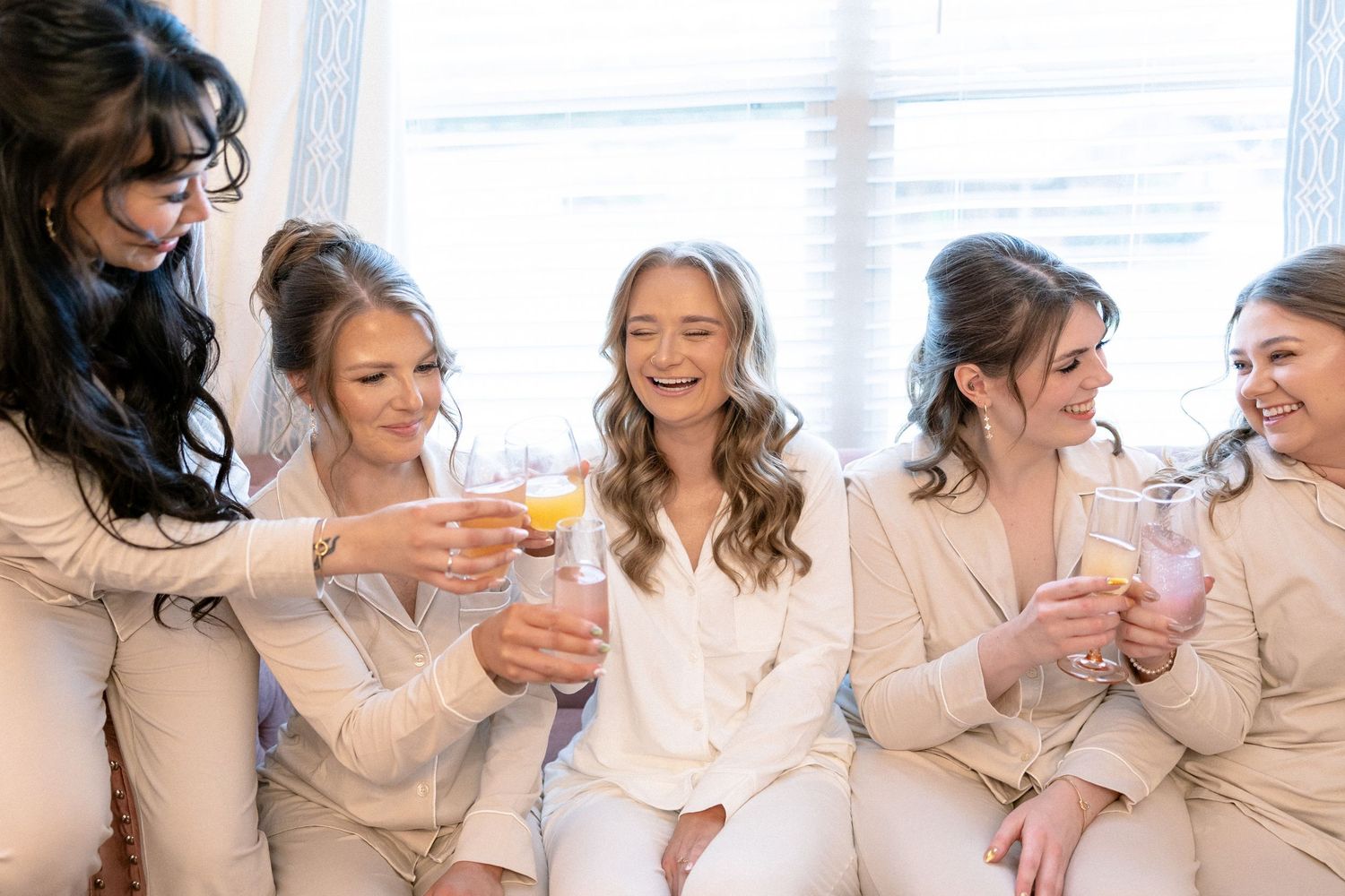 Bride and bridesmaids in cream satin pajamas toasting with mimosas before wedding.