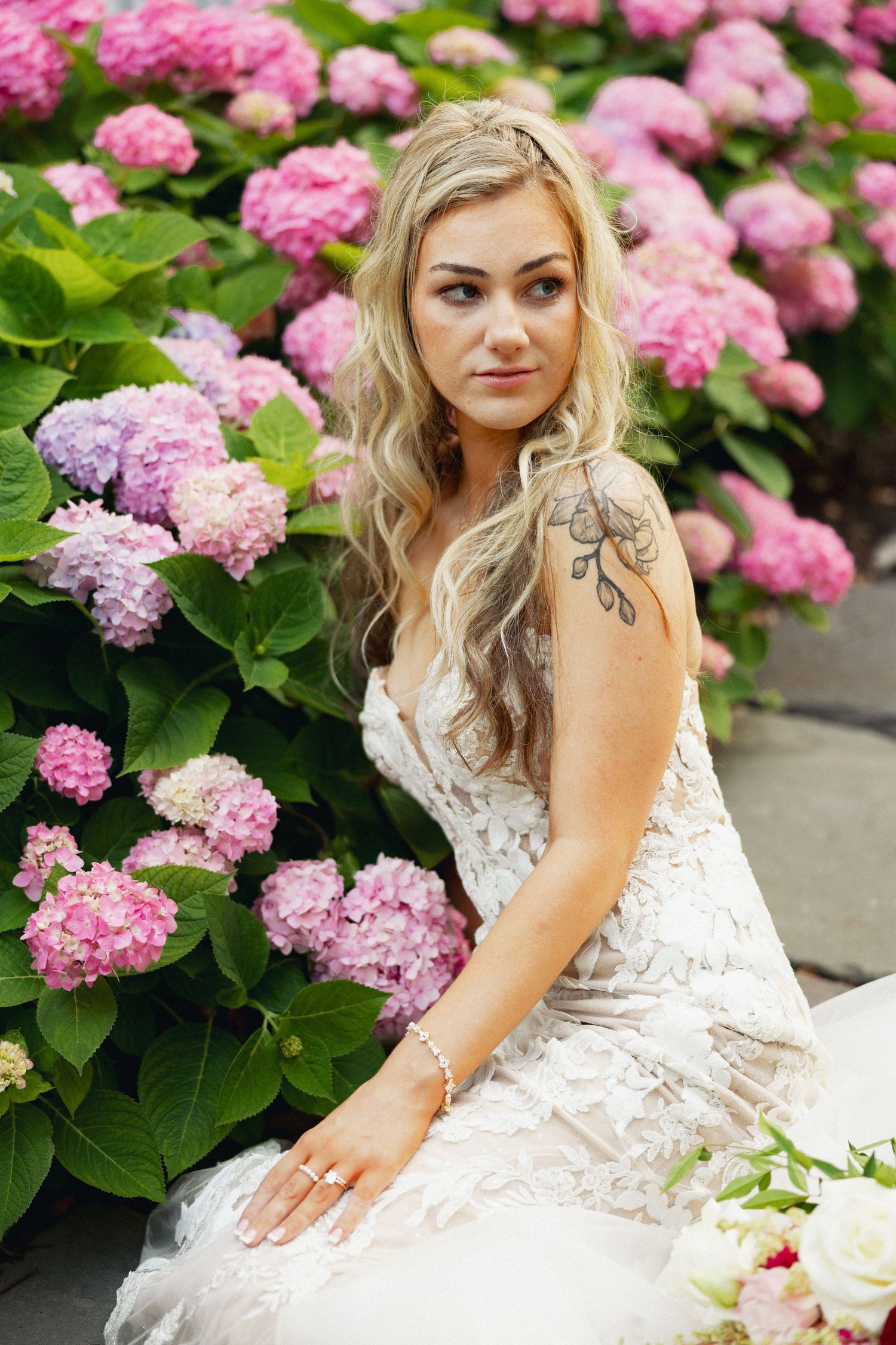 Blonde bride in lace wedding gown sitting among pink hydrangea blooms with floral arm tattoo.