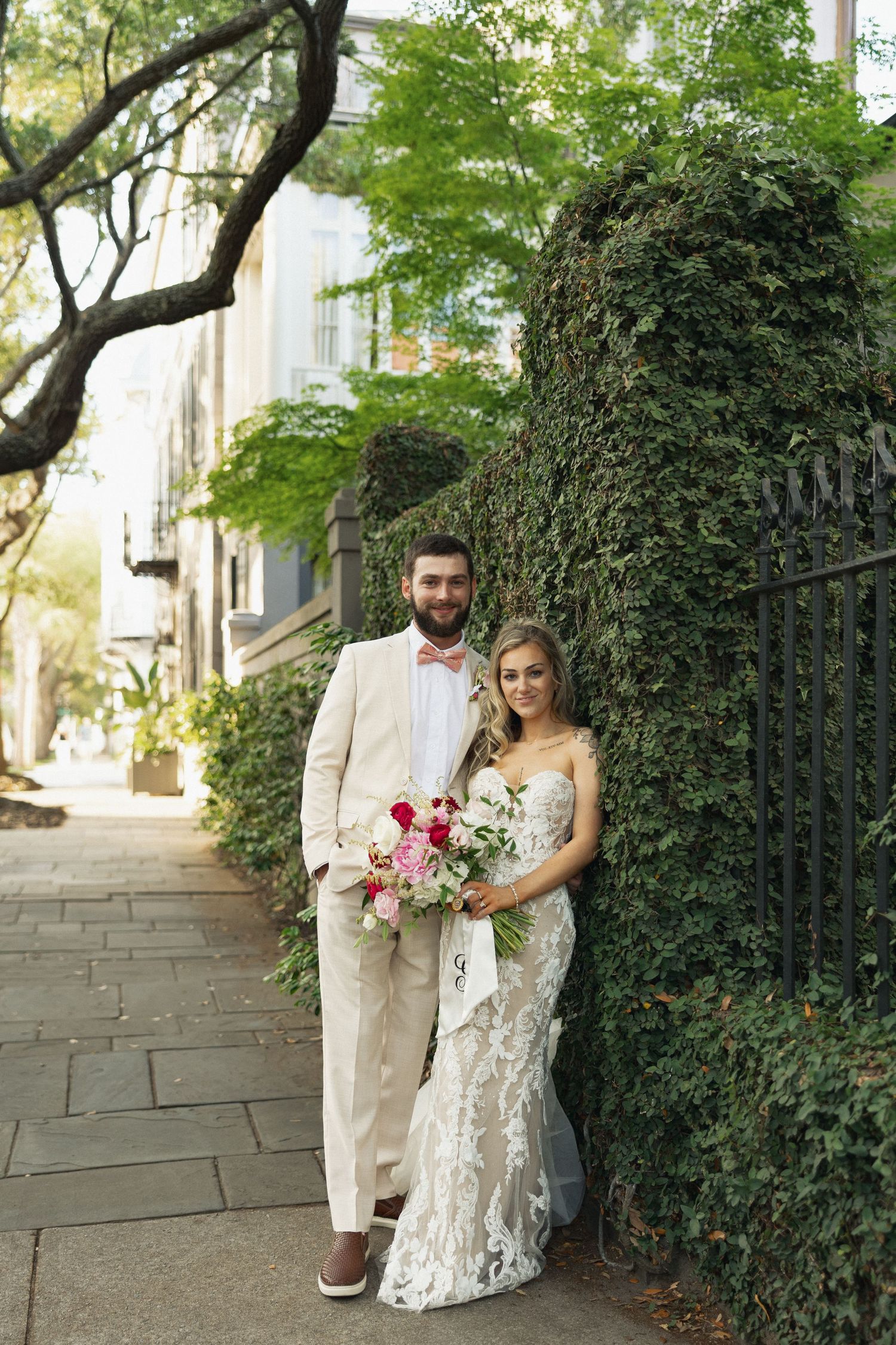Bride and groom pose by ivy-covered wall on Charleston sidewalk, she in lace gown holding pink bouquet, he in cream suit.