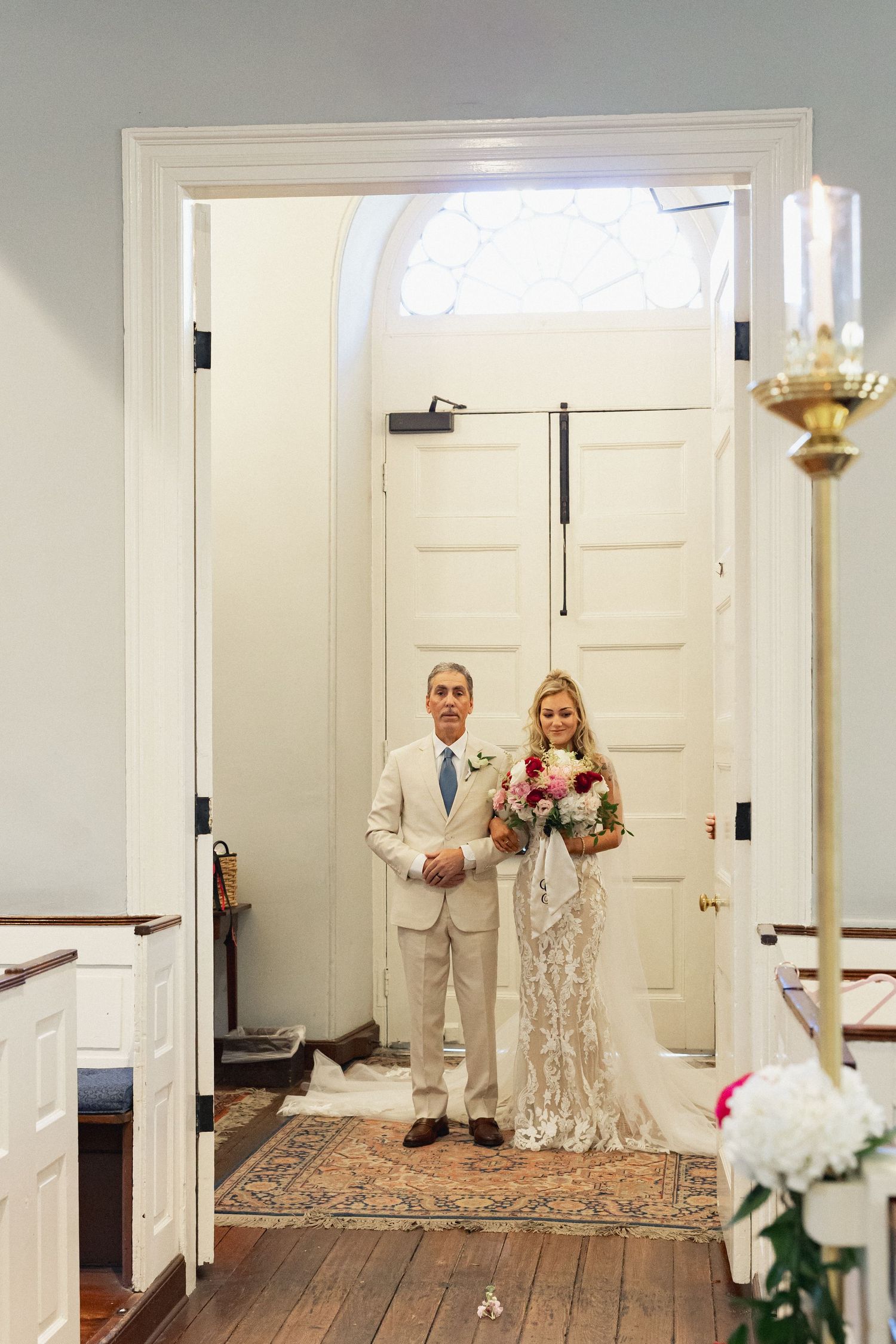 Bride in lace gown holds bouquet while standing with her father at church doors before walking the aisle.