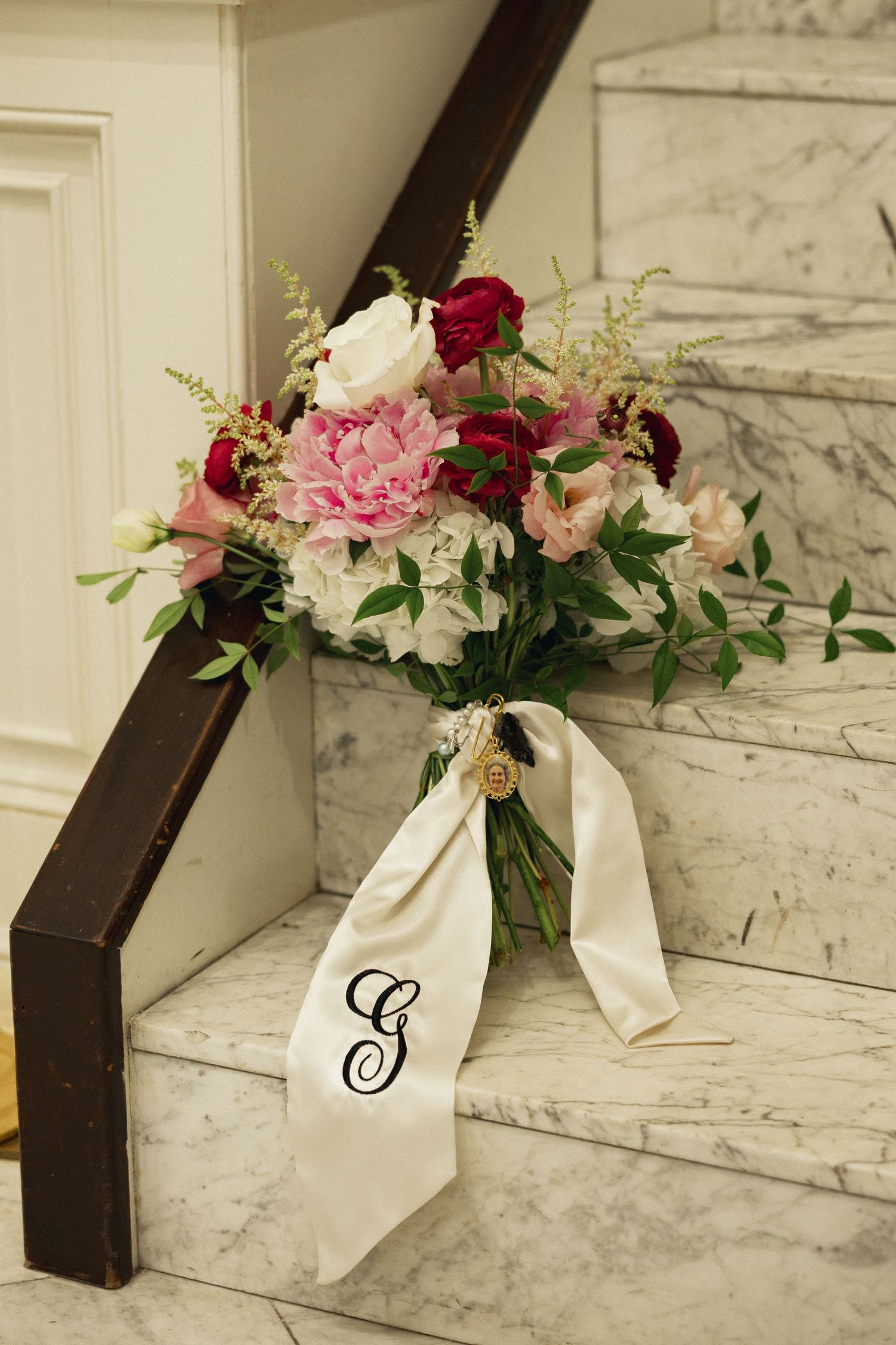 Bridal bouquet with pink, red, and white flowers tied with monogrammed ribbon on marble stairs.