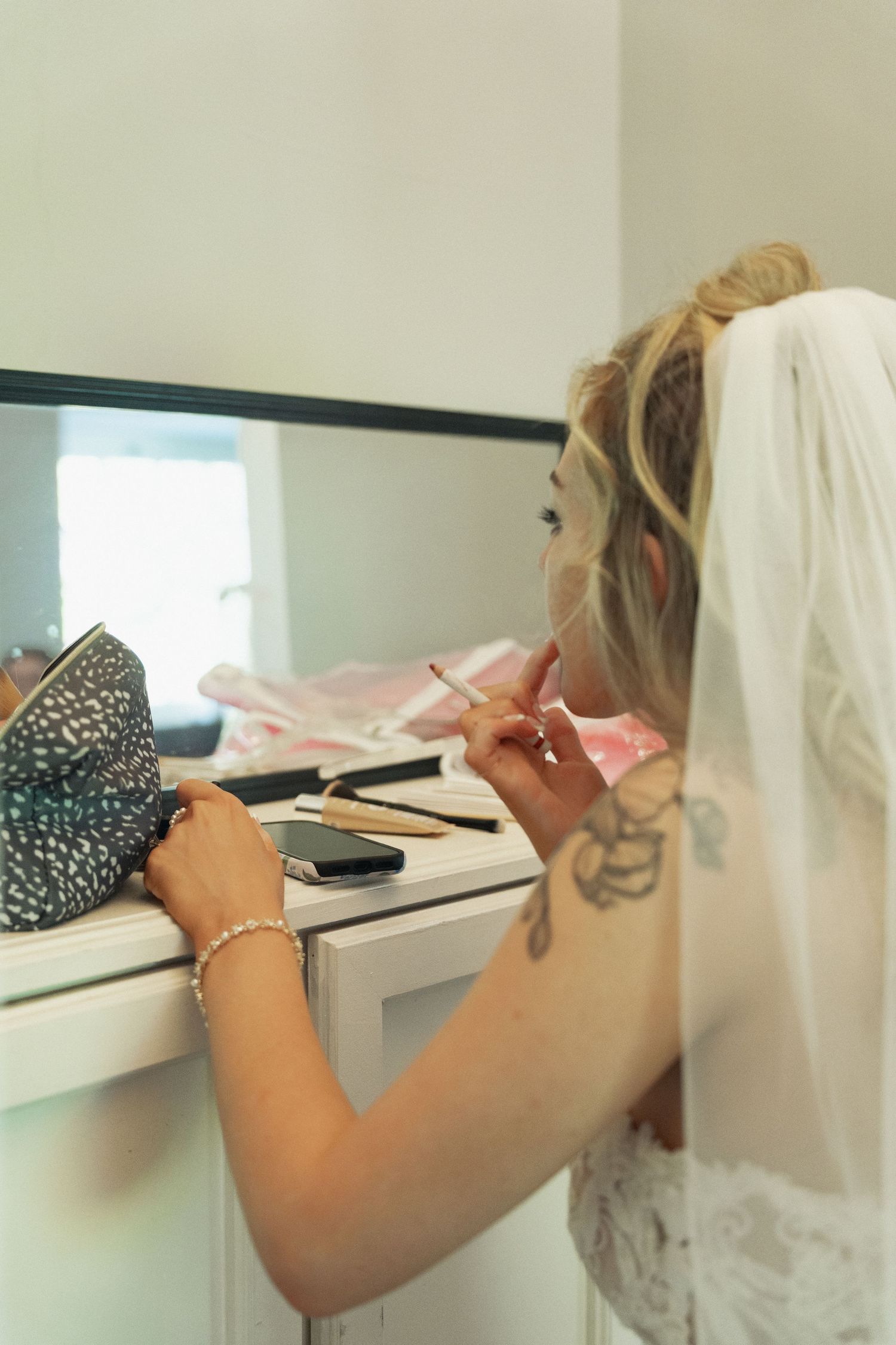 A bride wearing a white veil applies makeup at a vanity mirror before her wedding ceremony.