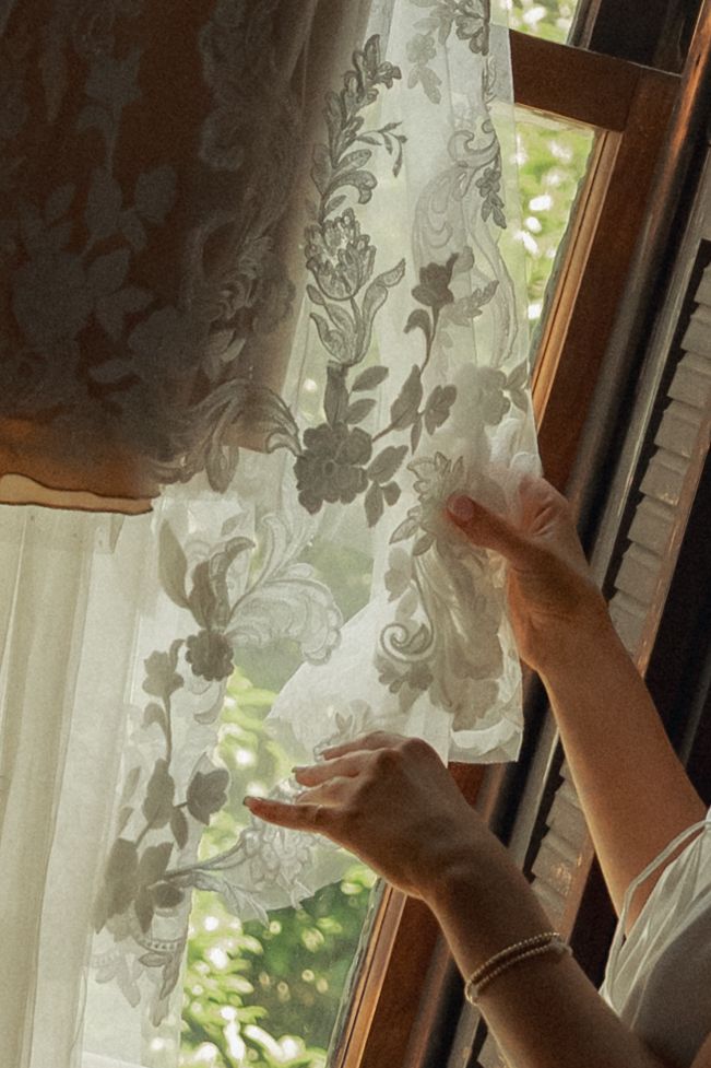 Hands gently pulling back sheer floral lace curtains on a wooden window frame with soft natural light filtering through.