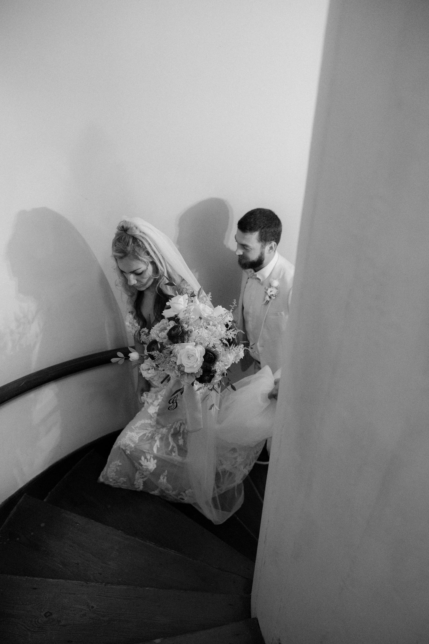 Black and white photo of bride and groom descending spiral staircase, bride holding bouquet in wedding gown.