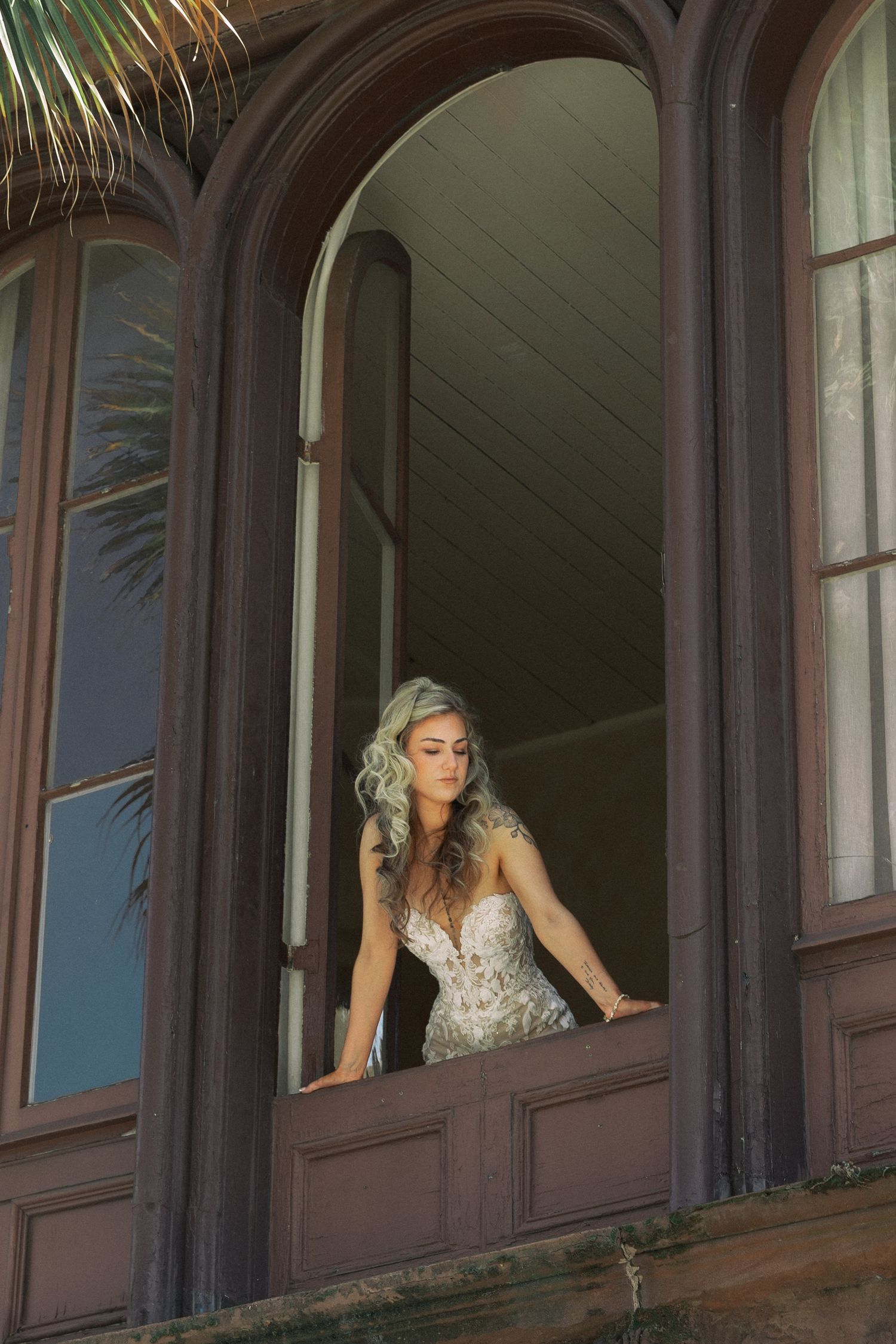 Bride in lace wedding gown standing in arched window of historic wooden building with palm trees.
