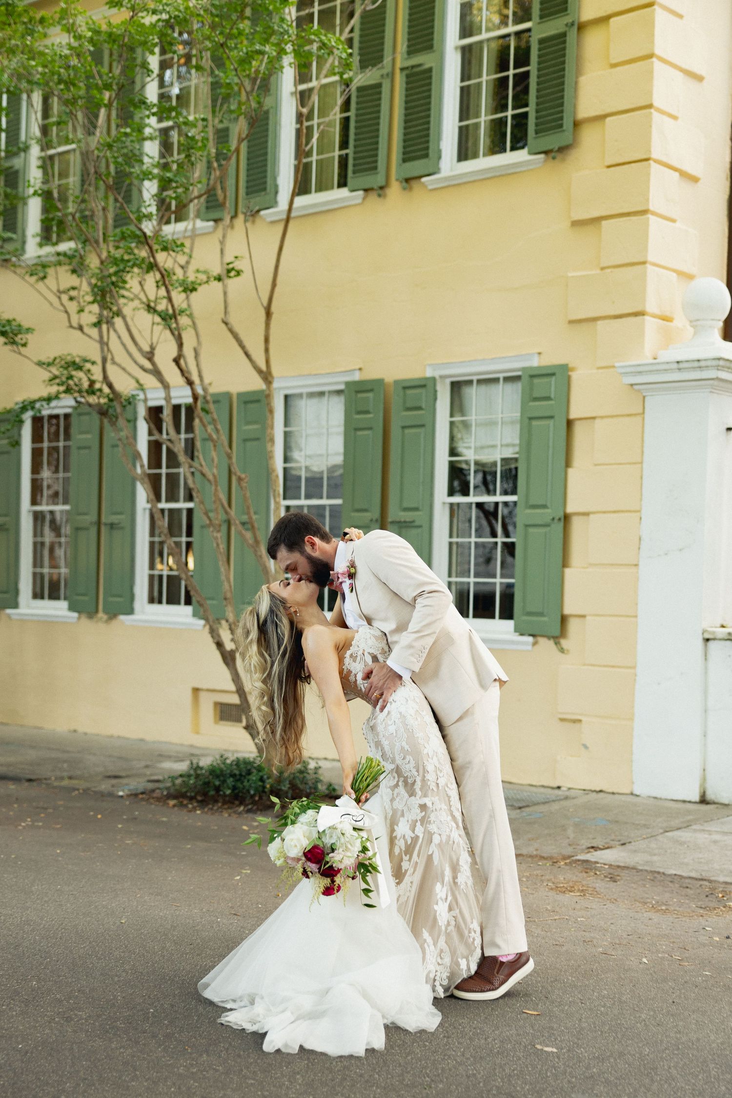 Groom dips bride for a kiss in front of yellow Charleston building with green shuttered windows, she holds bouquet.
