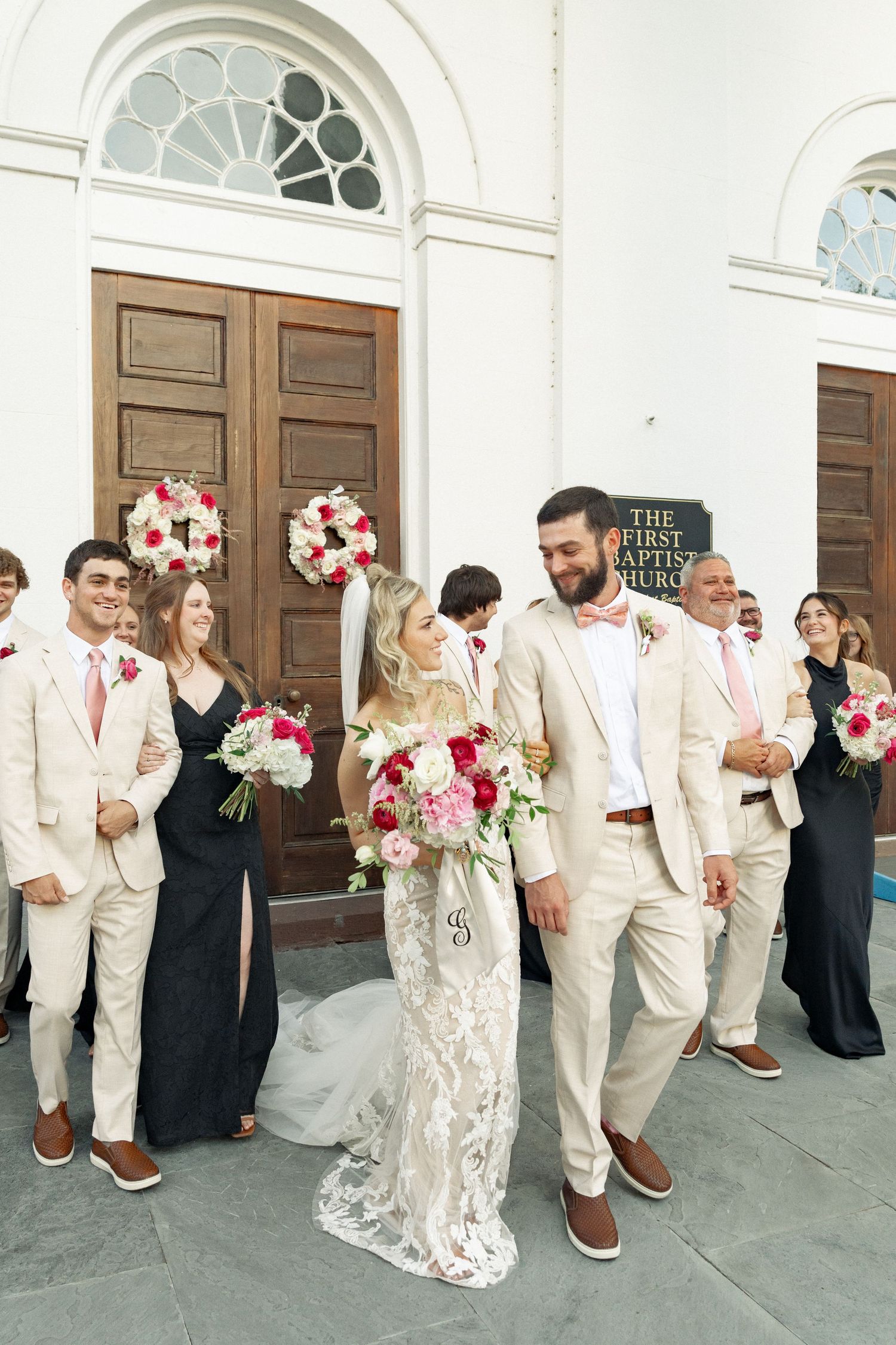 Bride and groom in tan suit walking with wedding party outside First Baptist Church on wedding day.