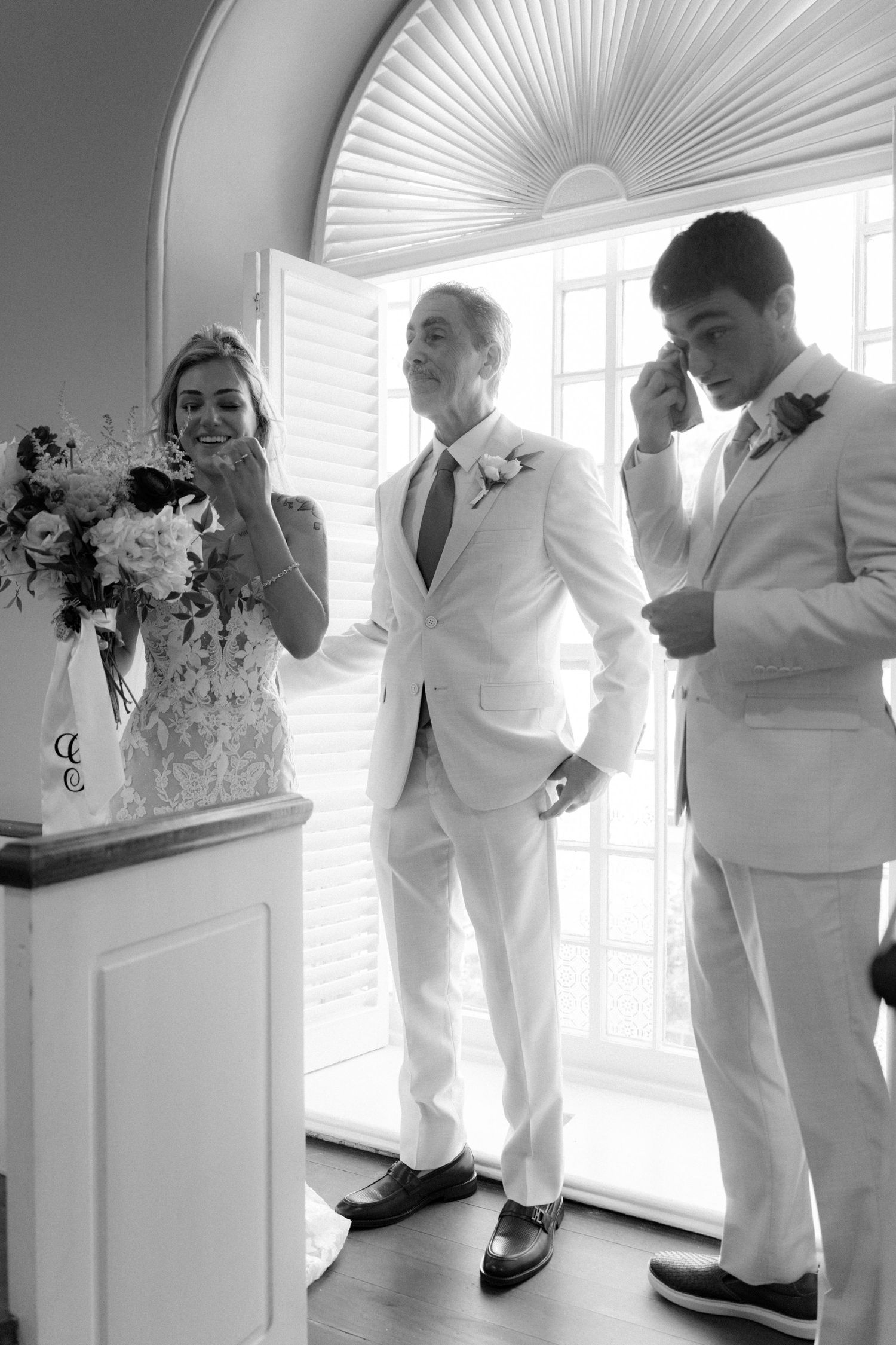Black and white photo of emotional bride, father, and groom during heartfelt wedding ceremony moment.