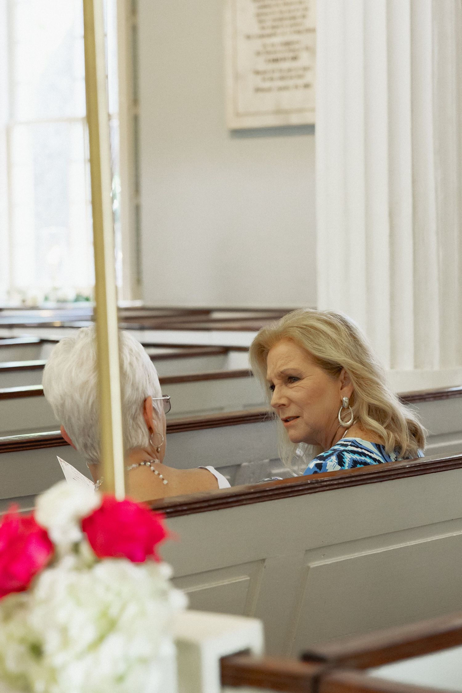 Two women conversing in a church pew during a wedding ceremony, with red and white floral arrangements in the foreground.