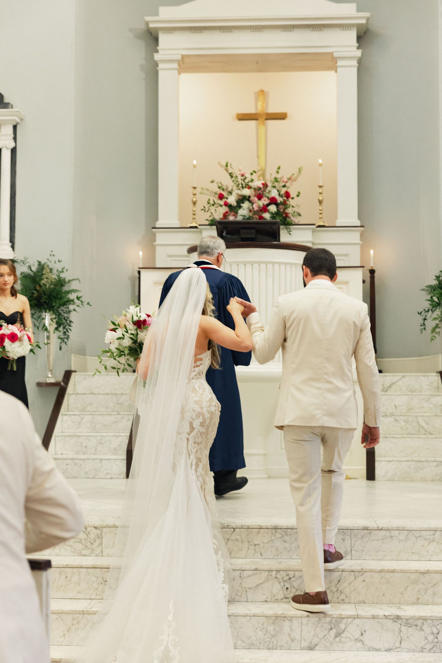 Bride in lace gown and groom in white suit ascend marble altar steps with officiant during ceremony.