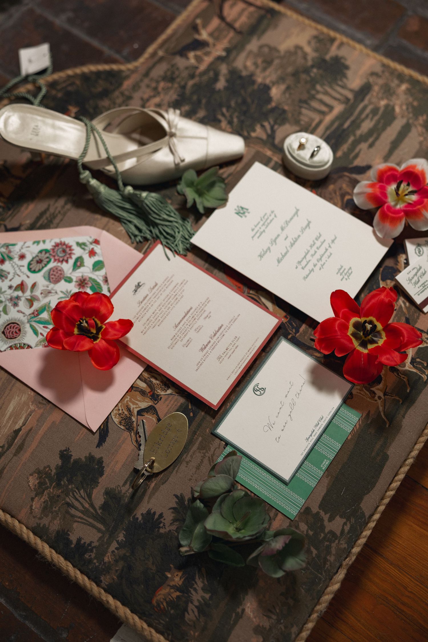 Wedding invitation suite with red tulips, pink envelope, and bridal shoes on vintage tray.