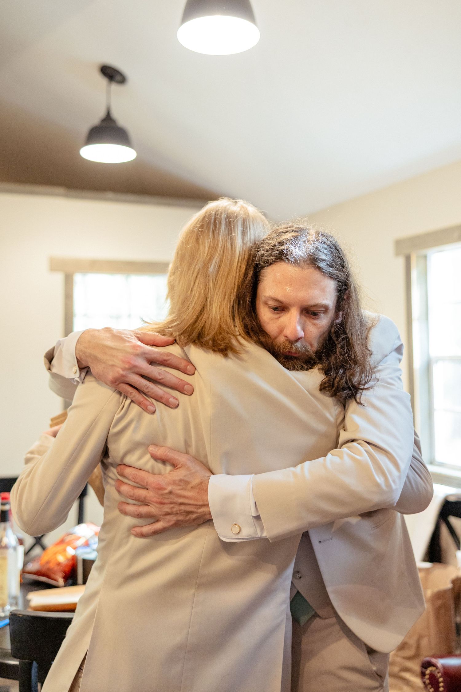 Long-haired groomsman in beige suit shares a heartfelt hug with a blonde woman before the wedding.