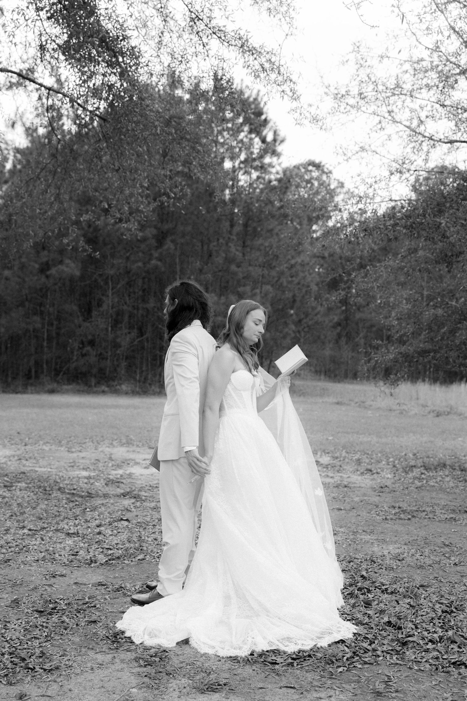 Black and white photo of bride and groom standing back-to-back reading vows in wooded setting.