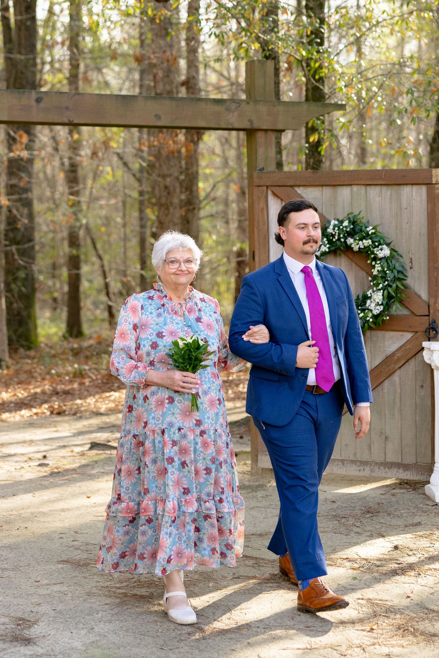 Elderly woman in floral dress holding white flowers being escorted by a young man in a navy suit at a wedding.