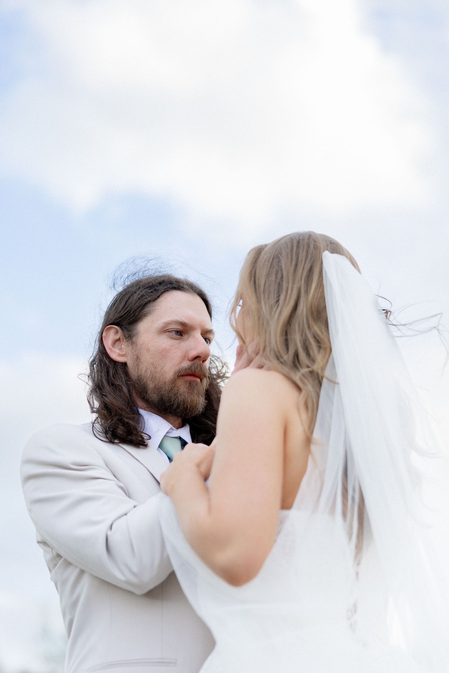 Bearded groom in gray suit gazes at bride in white veil against blue sky.
