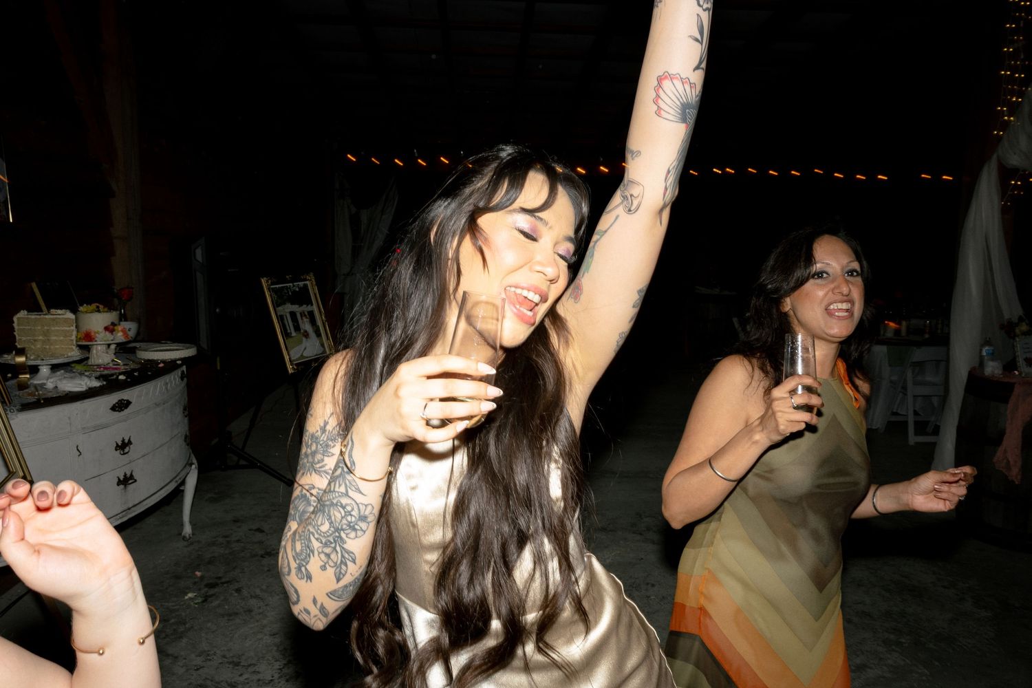Tattooed woman in gold dress dances energetically with arm raised at a wedding reception.