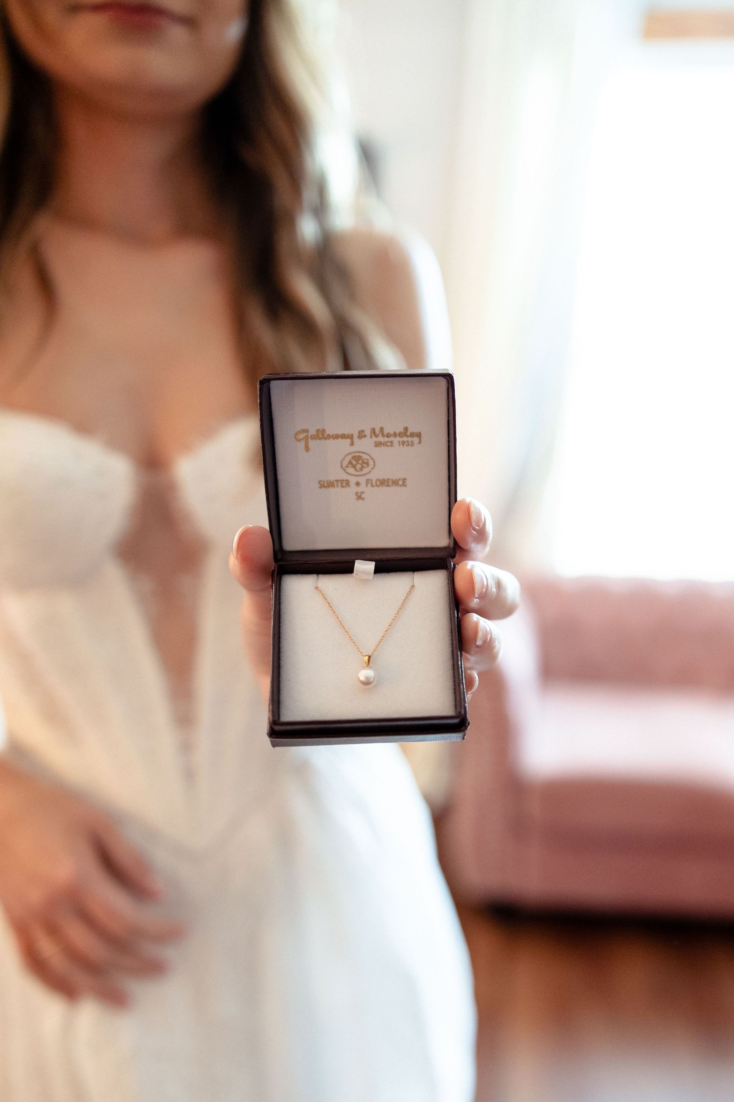Bride holding open a jewelry box displaying a delicate gold pearl pendant necklace on her wedding day.