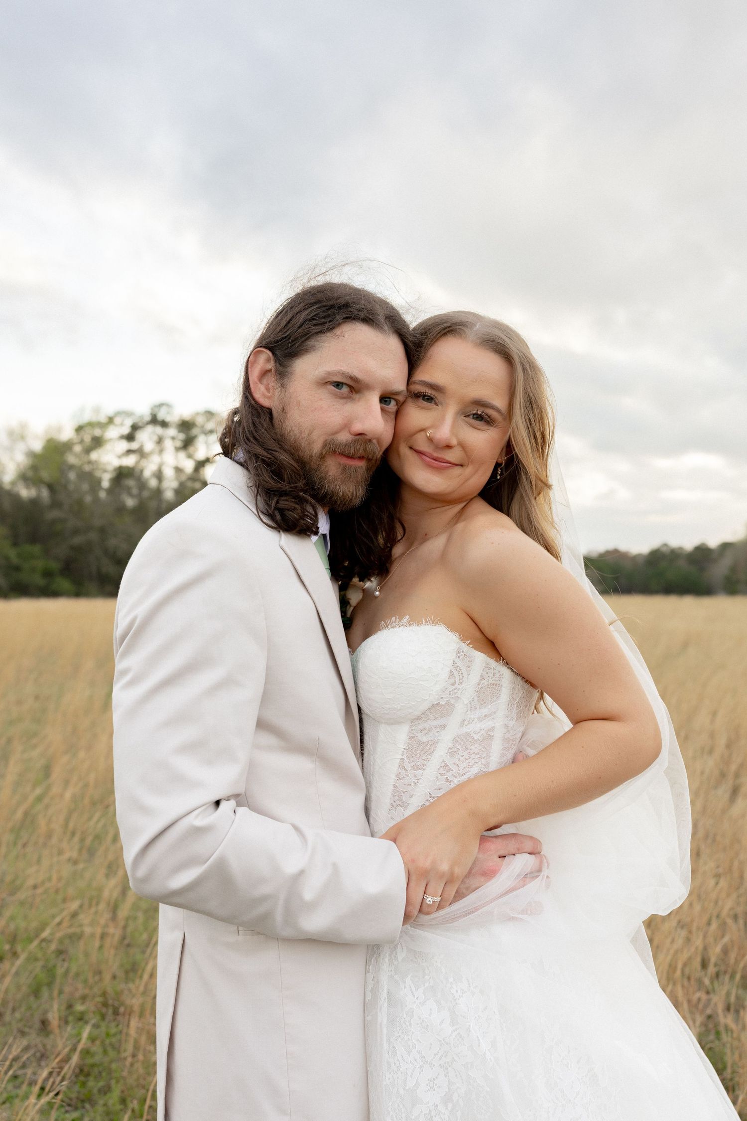 Bride and groom embrace in golden field, she wears white lace gown with veil, he wears ivory suit, cloudy sky behind.