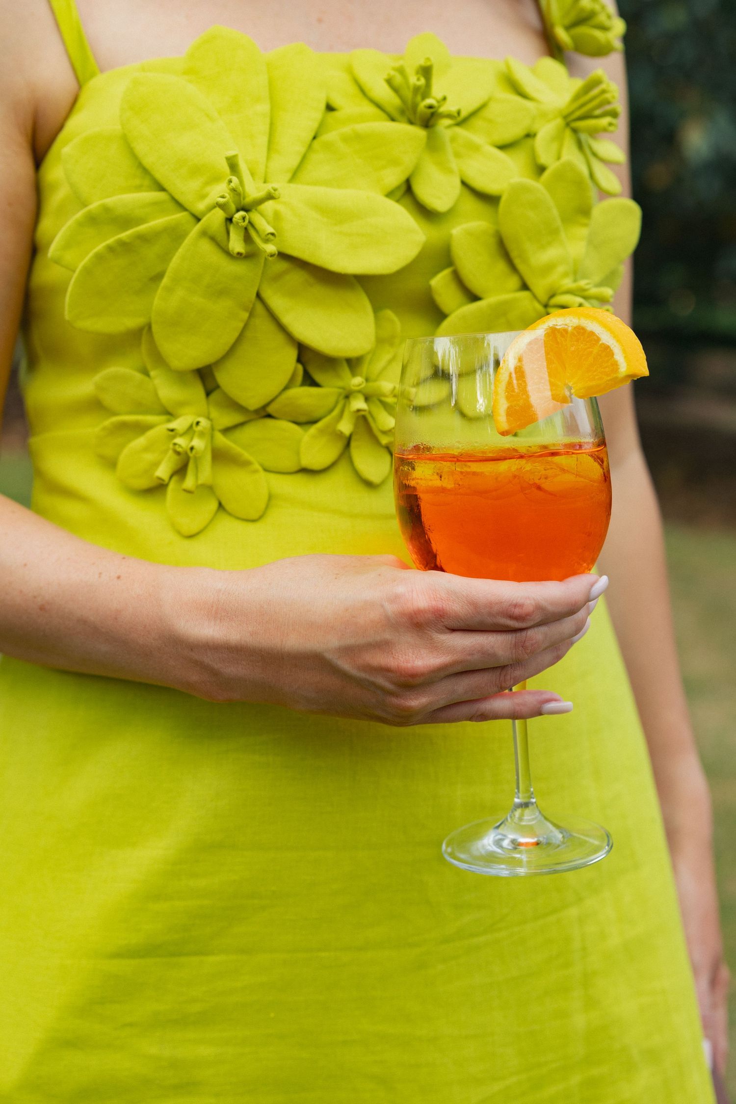 Woman in yellow-green floral dress holding an Aperol Spritz cocktail garnished with an orange slice.