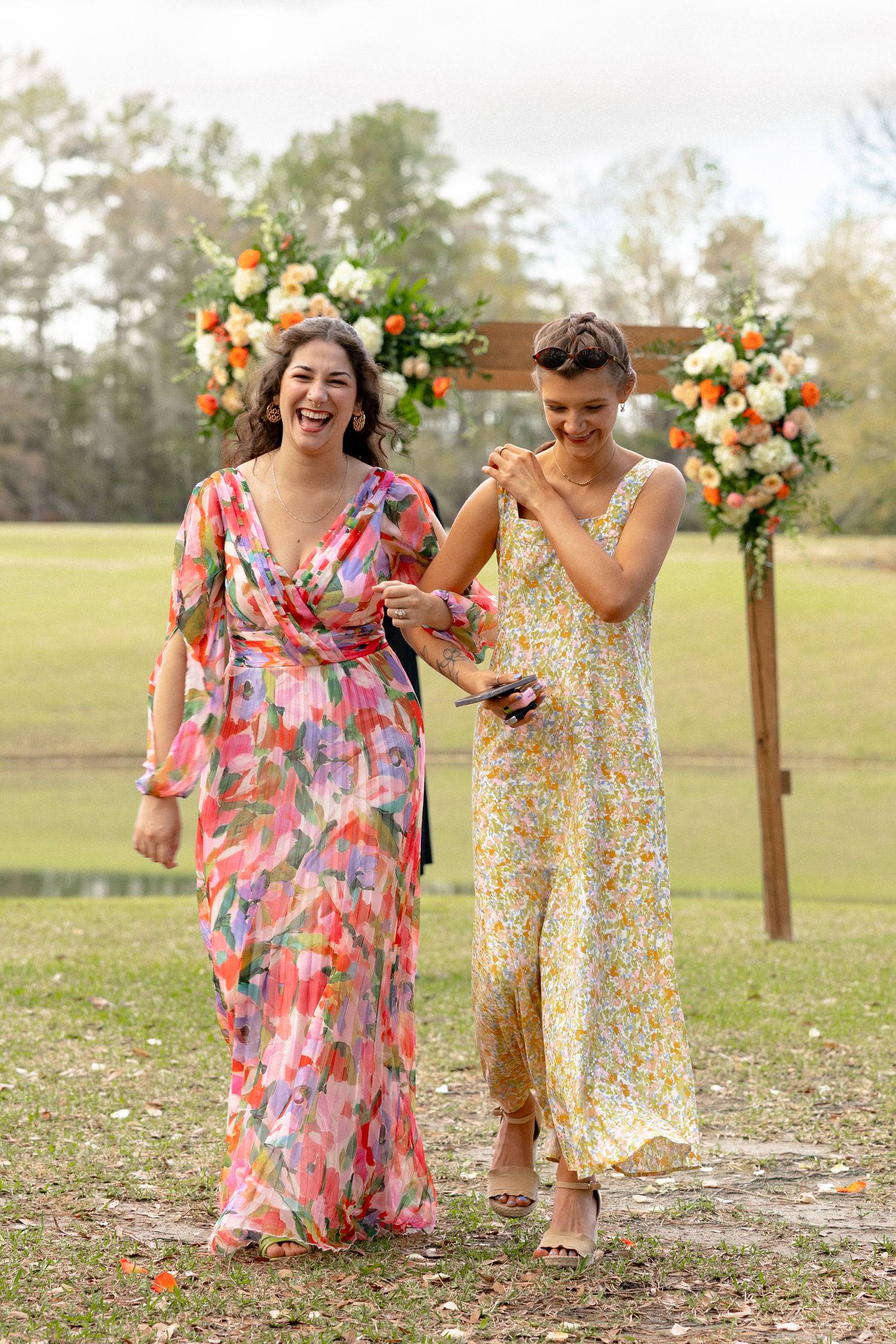 Two women in colorful floral dresses laugh joyfully in front of wooden wedding arch with flowers.