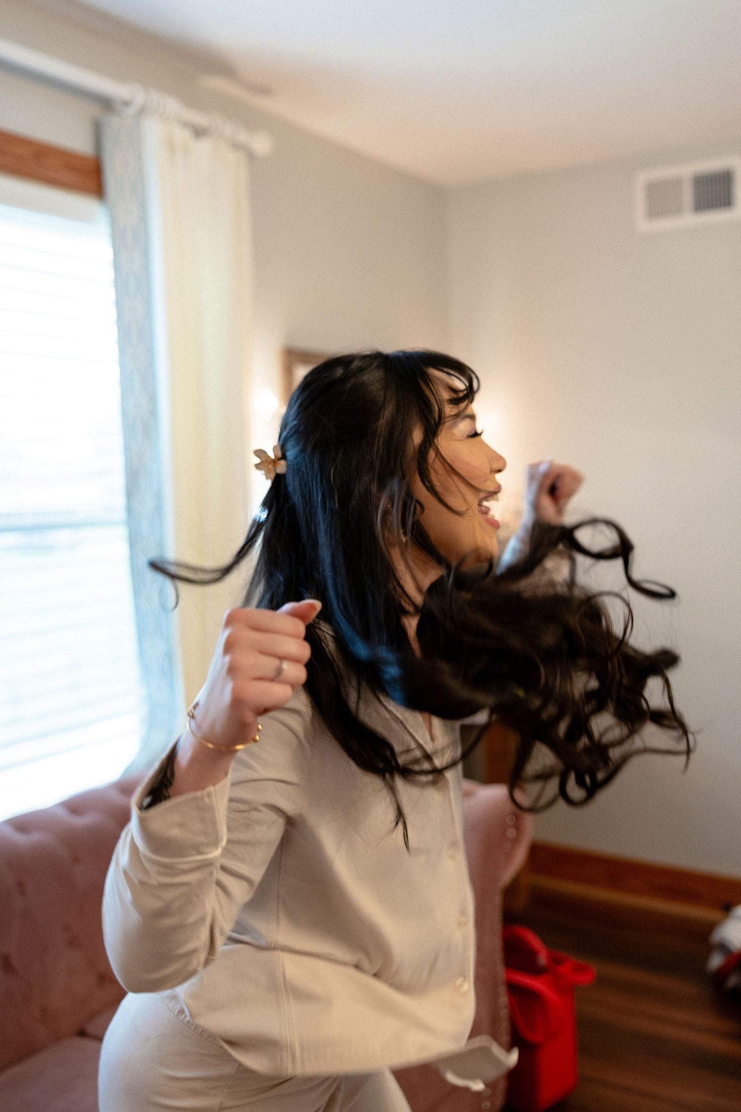 Brunette bridesmaid flipping long curly hair extensions in bright bedroom.