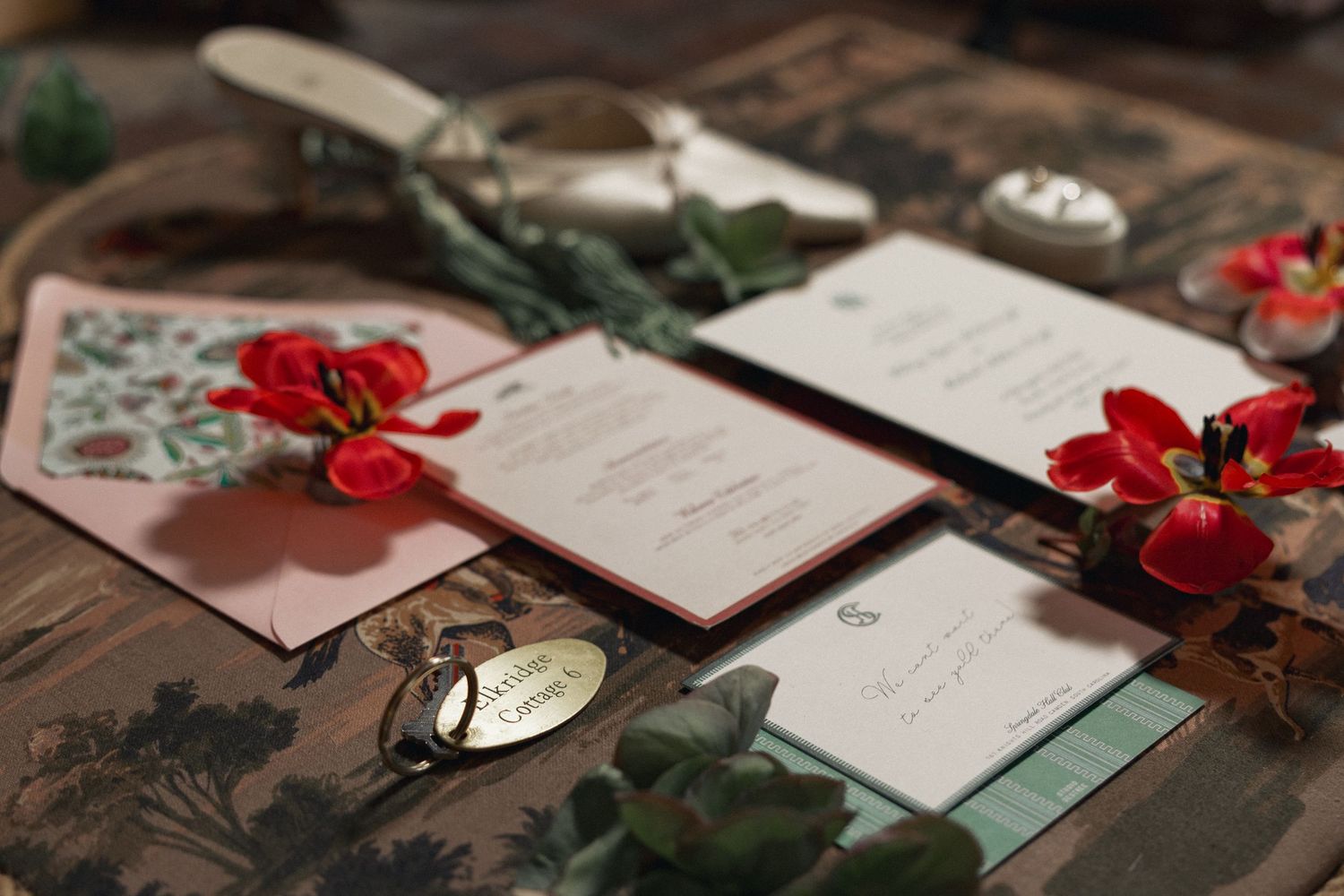 Elegant wedding stationery suite with red floral accents, pink envelope, and greenery on a rustic wooden surface.