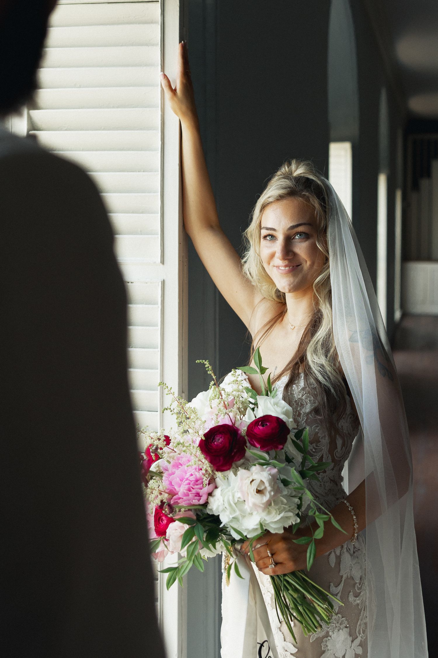 Smiling blonde bride in lace gown holding red and white peony bouquet, wearing veil, posing by window on wedding day.